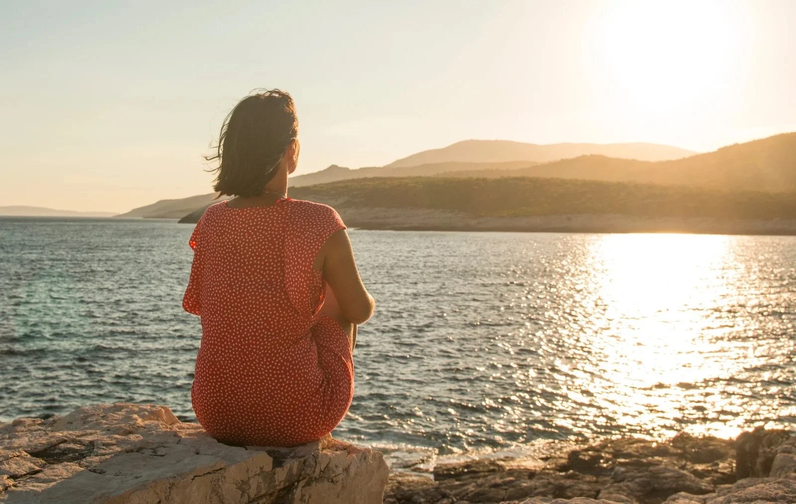 Woman sitting on rocky shore, facing water and mountains during sunset.
