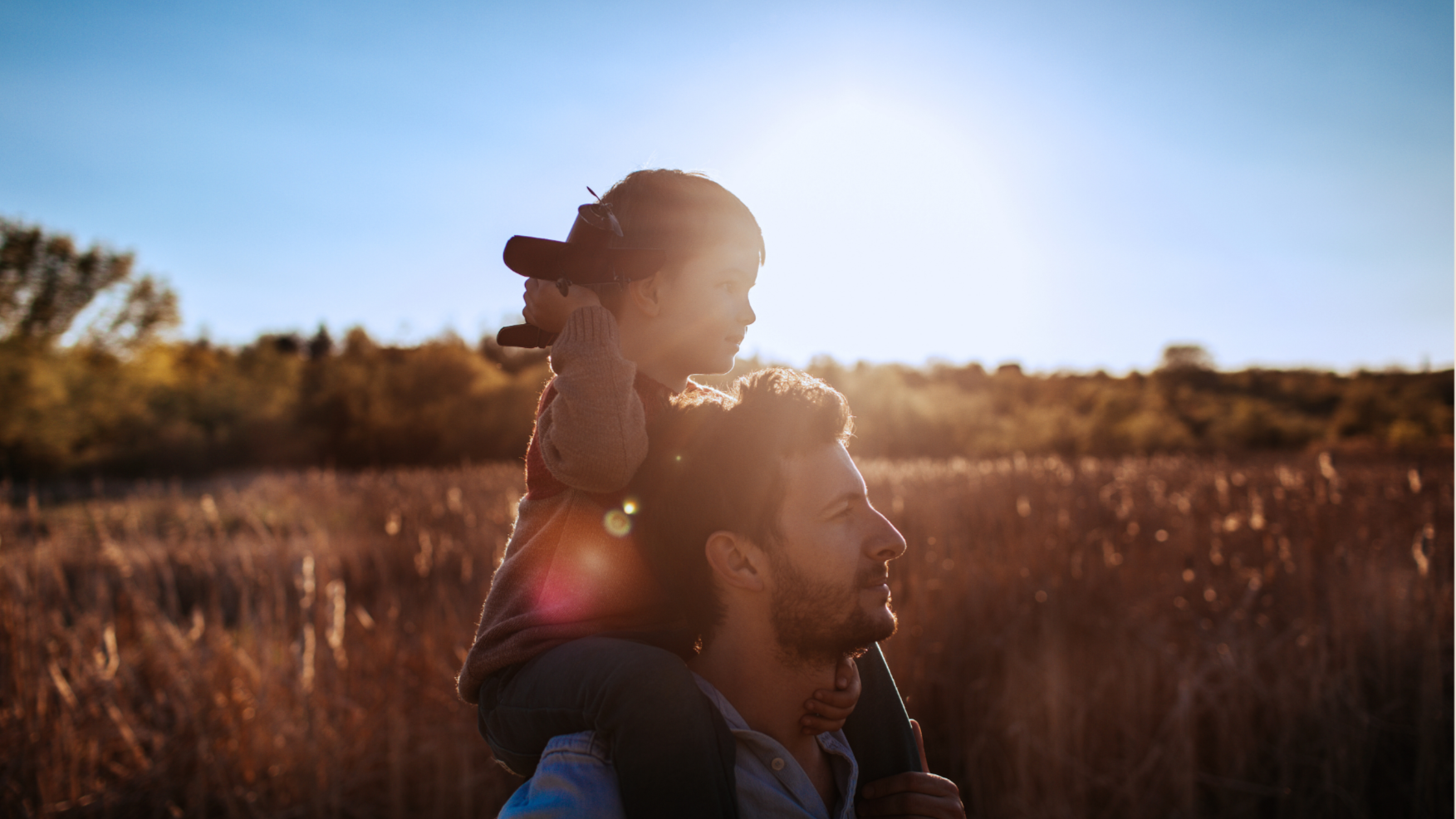A father carrying his young child on his shoulders in a field during sunset, with the sun behind them.