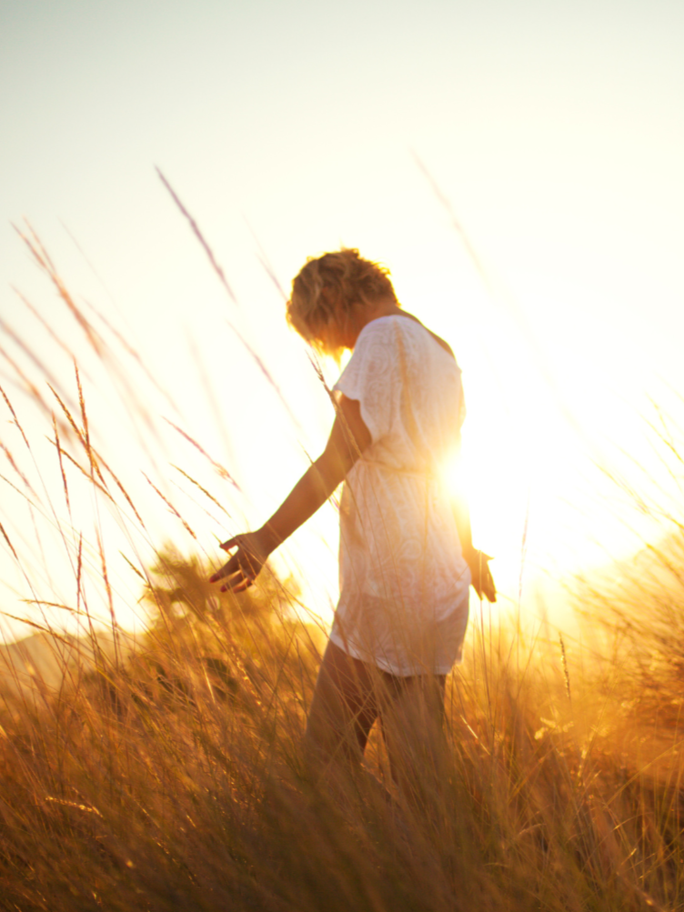A person with blonde hair wearing a white dress walking through a field of tall grass during sunset.