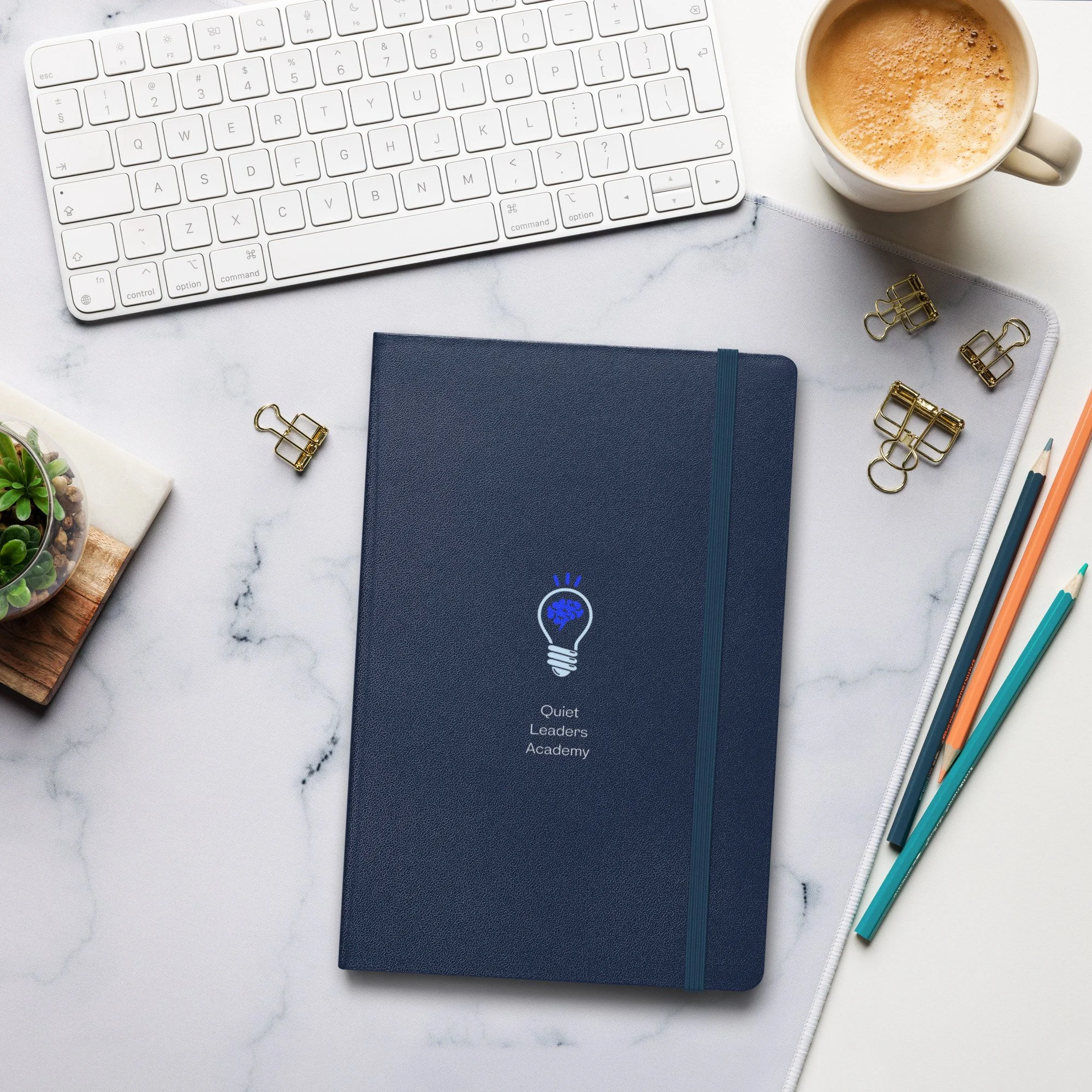 A flat lay of a workspace with a white keyboard, a cup of coffee, a closed navy blue notebook with 'Quiet Leaders Academy' and a lightbulb logo, several gold binder clips, a set of colored pencils, a small potted succulent, and a white desk mat.