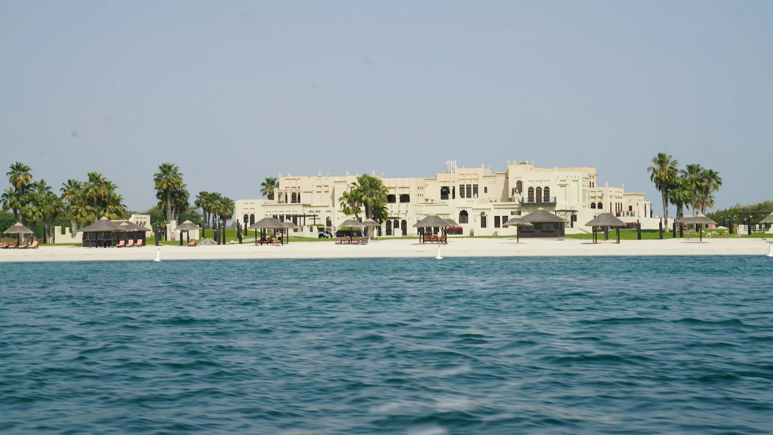 Luxury resort or hotel on a waterside with palm trees, umbrellas, and beach chairs, overlooking blue water under a clear sky.