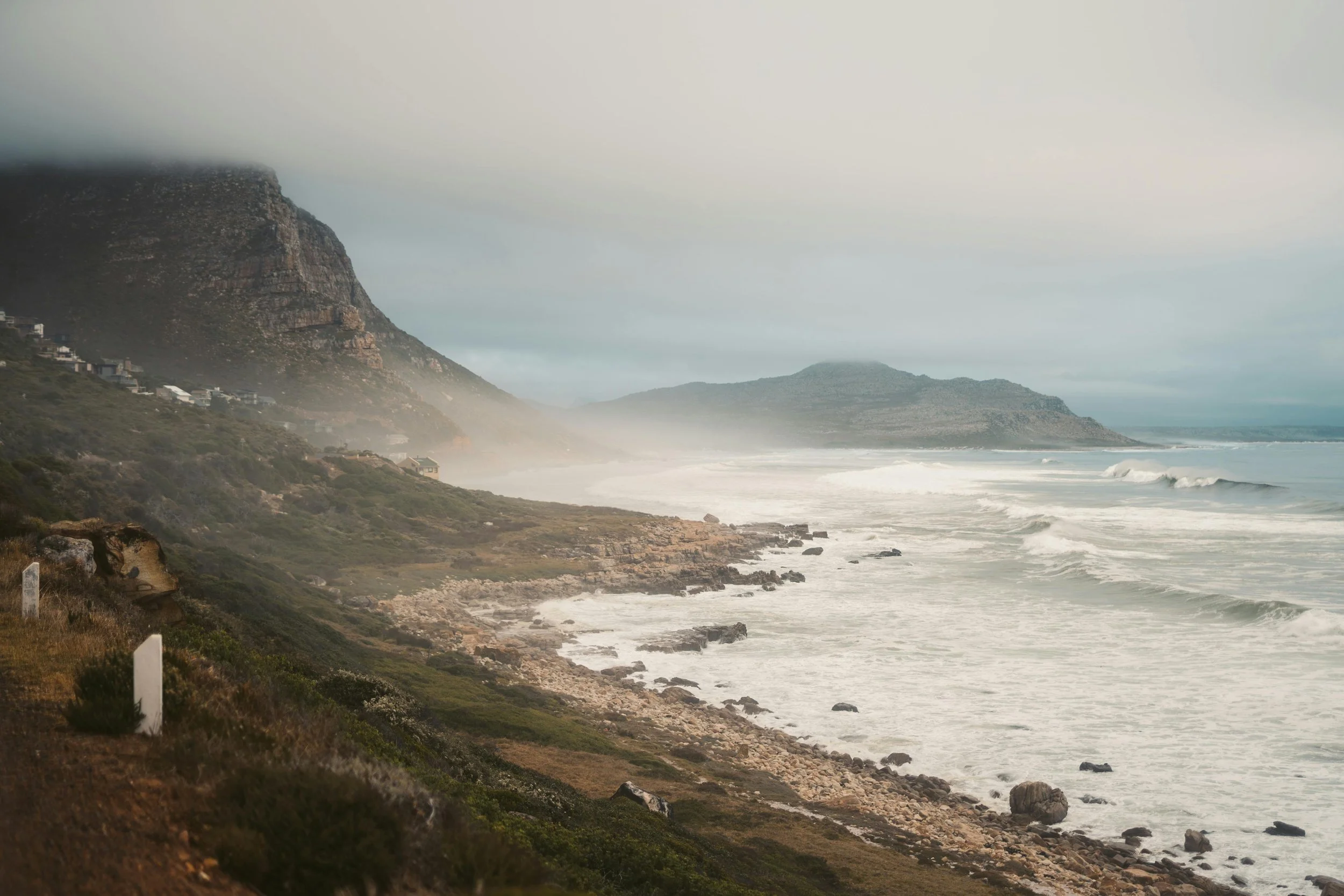 Misty Cape coastline with waves breaking along rocky shores beneath mountain slopes.