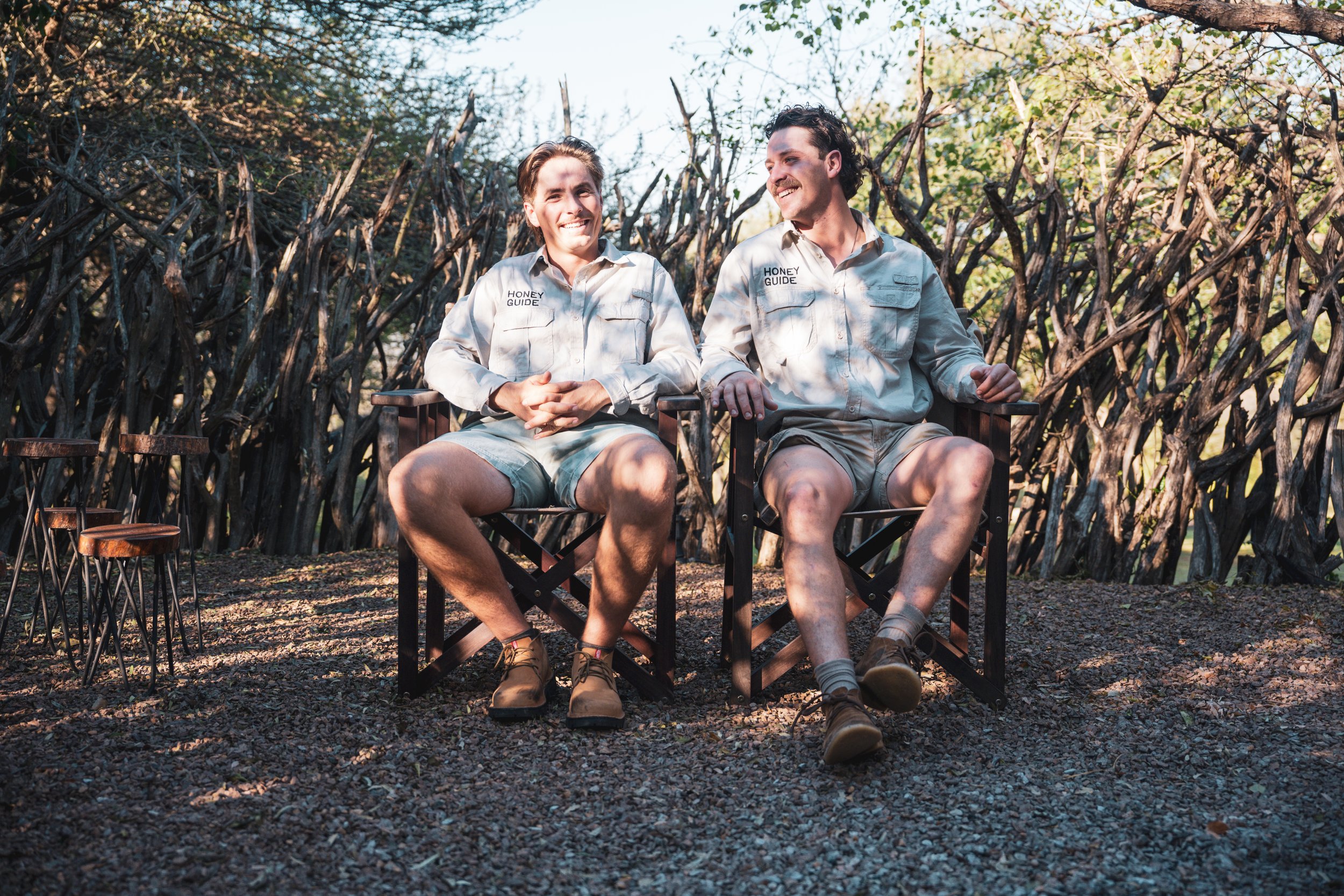 Two men sitting outdoors on black chairs, smiling and talking, with a background of bare branches and trees.