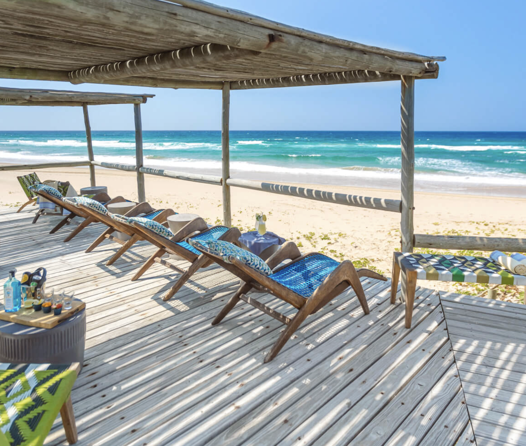 Beach scene with wooden lounge chairs under a canopy, white sand, and ocean waves under a clear blue sky.