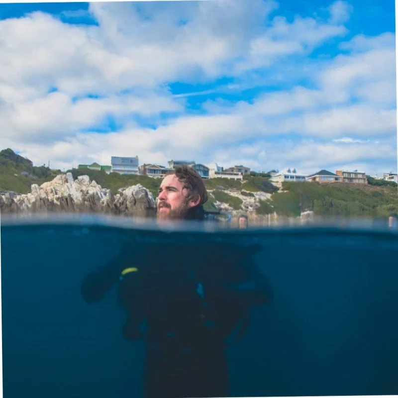 A person partially submerged in water, with only the head above the surface, and houses on a hillside in the background under a partly cloudy sky.