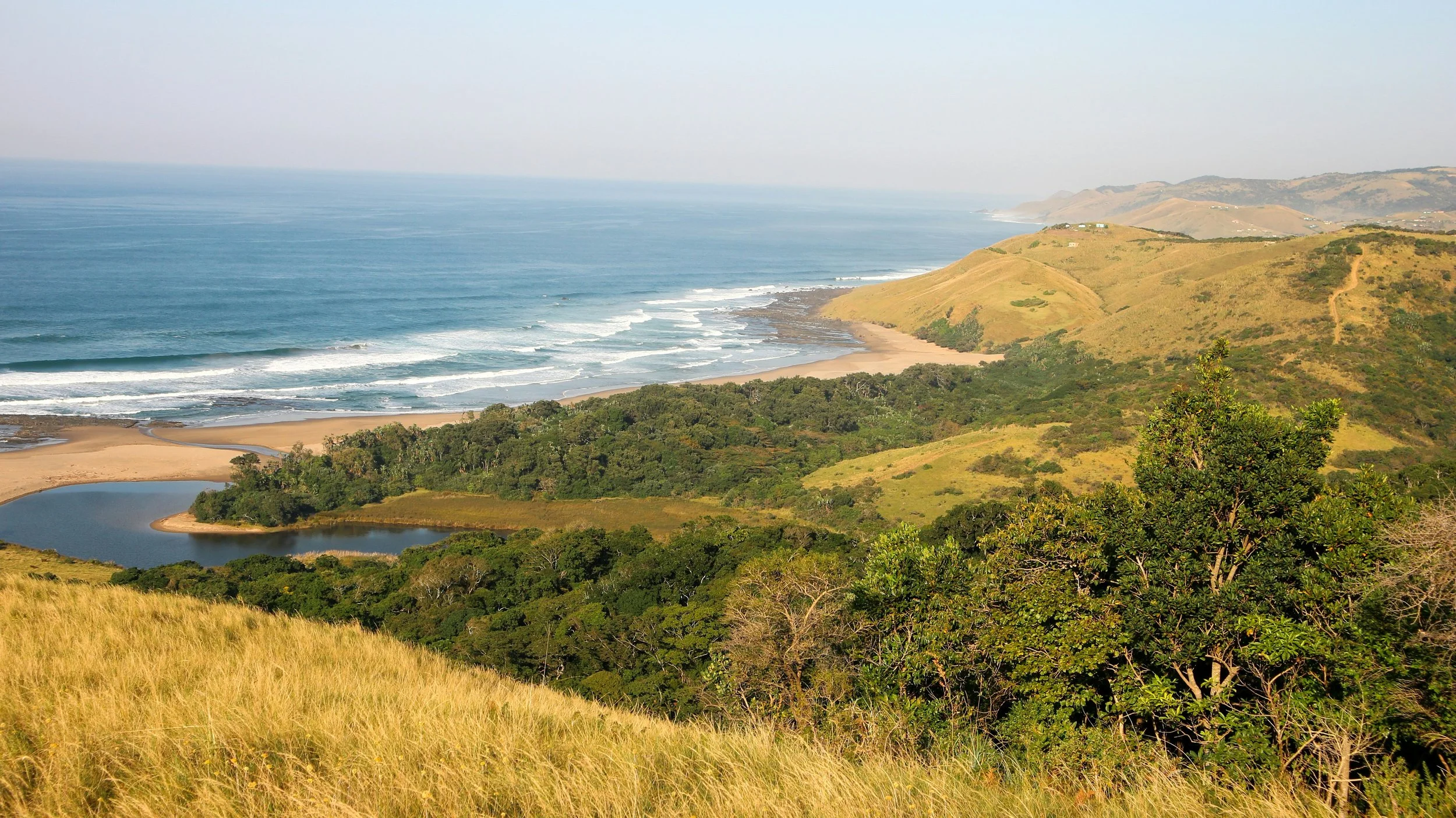 Scenic view of a coastal landscape with rolling green hills, a beach with waves, a small pond, and lush vegetation under a clear sky.