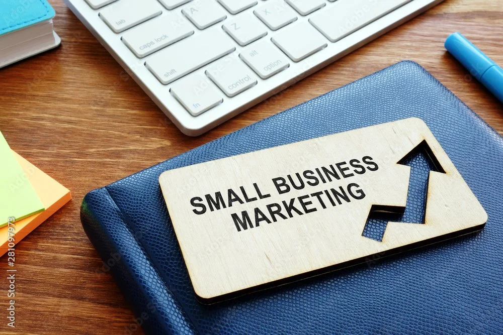 Wooden sign with the words 'Small Business Marketing' and an upward trending arrow. Office desk with keyboard, blue pen, colorful sticky notes, and notebook.