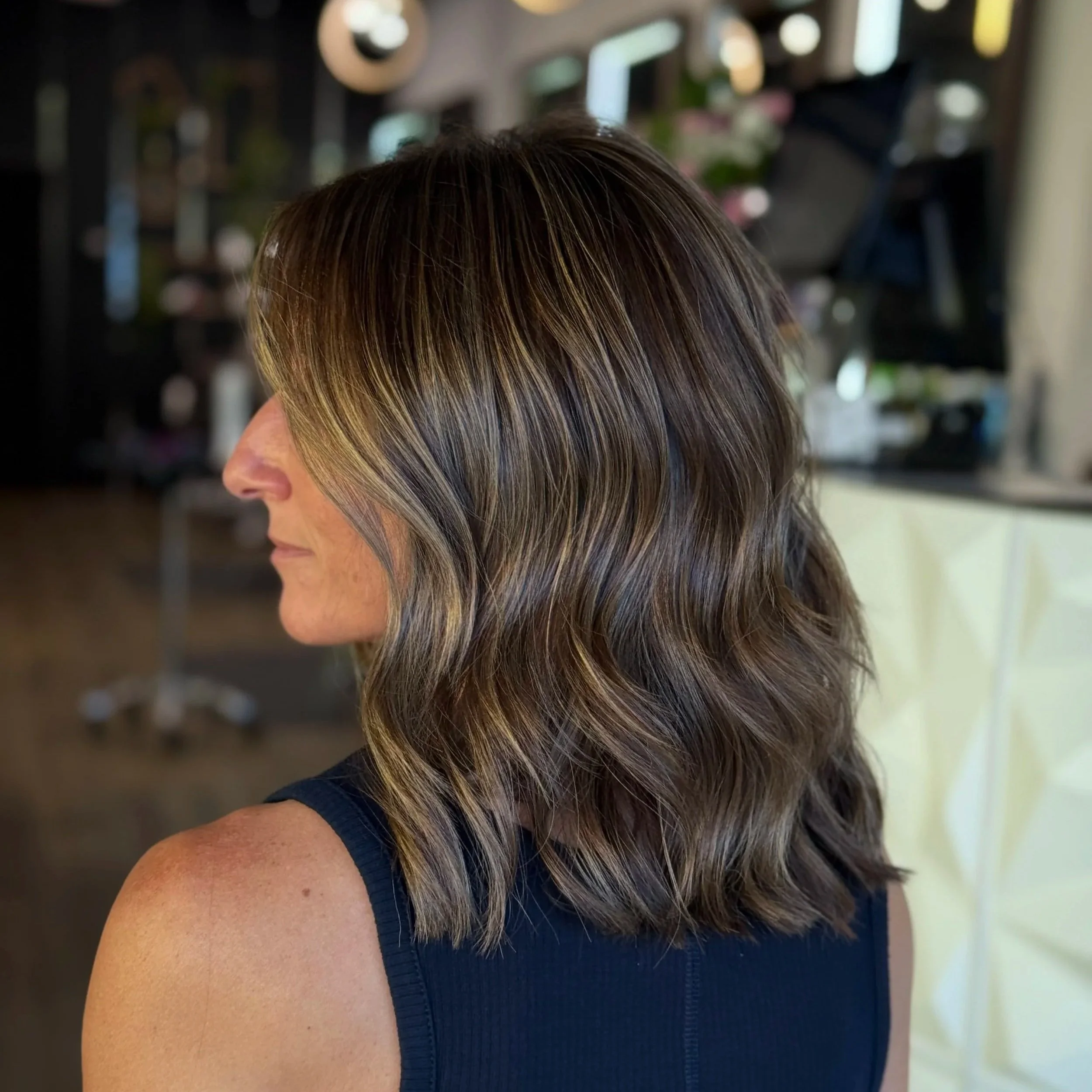 Side view of a woman with shoulder-length wavy brown hair with blonde highlights, wearing a navy sleeveless top, indoors with blurred background.
