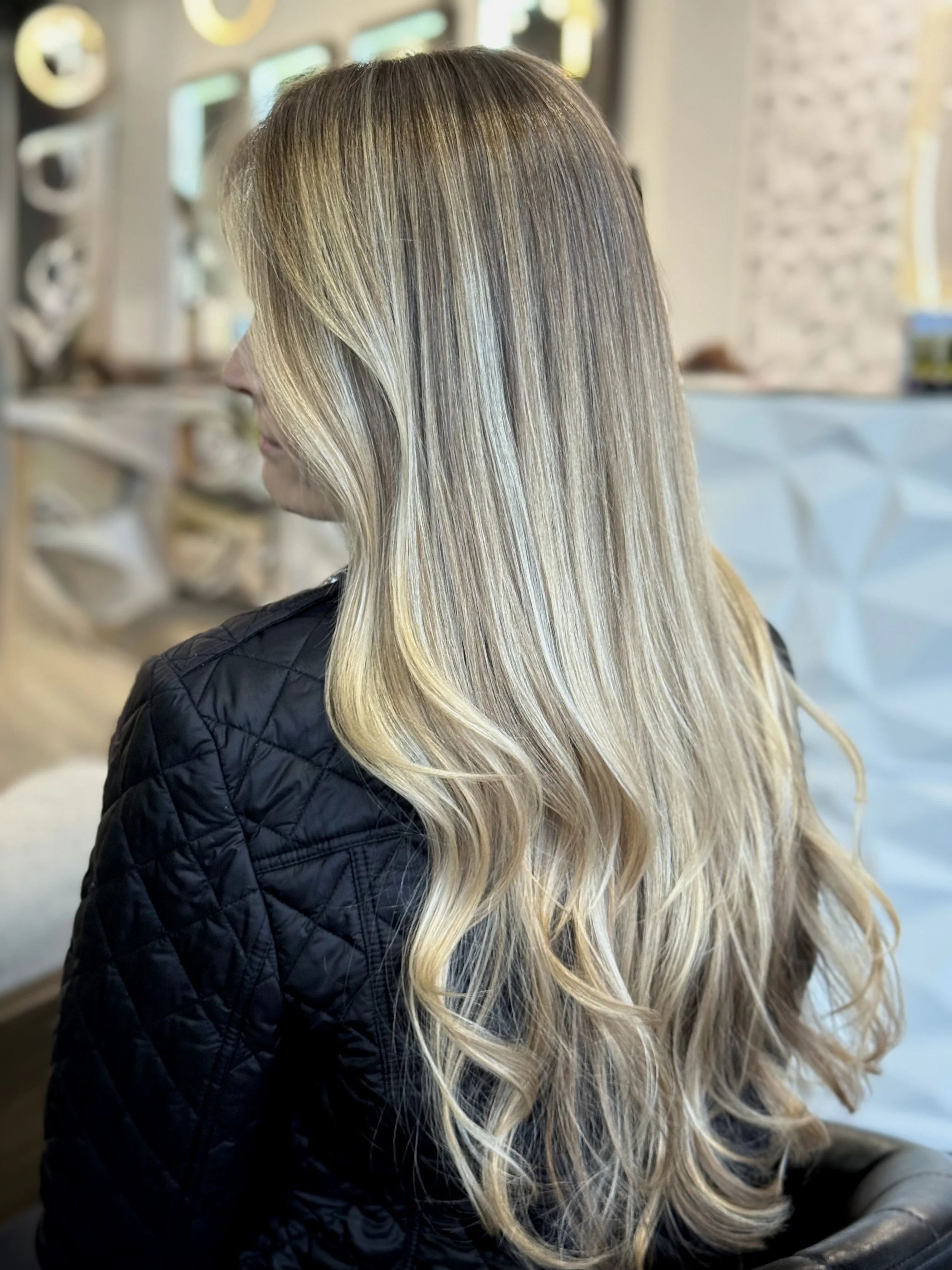Woman with long, wavy blonde hair sitting in a salon or studio with modern decor.