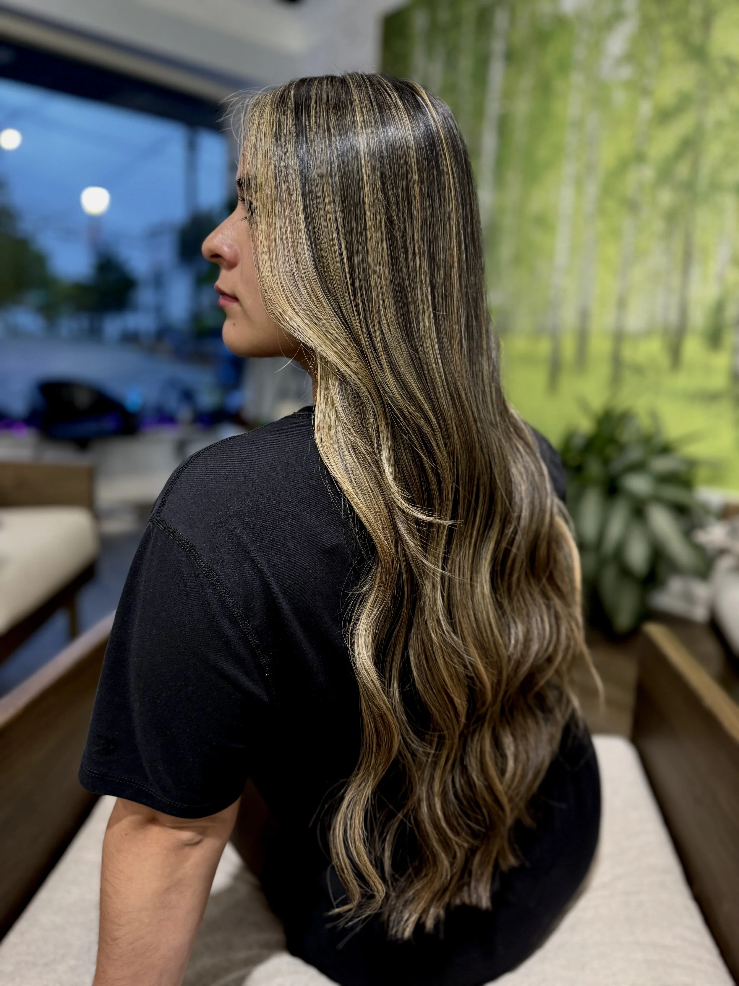 Side view of a woman with long, wavy hair with highlights, sitting indoors with a green forest mural in the background.