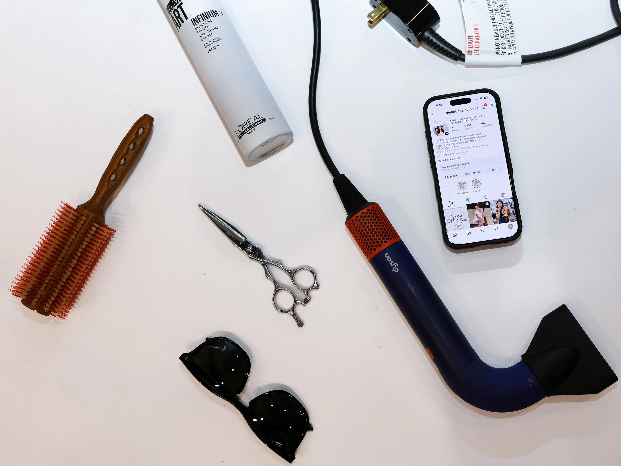 A white surface with a hairbrush, small scissors, sunglasses, a tube of L'Oreal hair product, a smartphone displaying a social media profile, a black plug, and a red and blue curling iron.