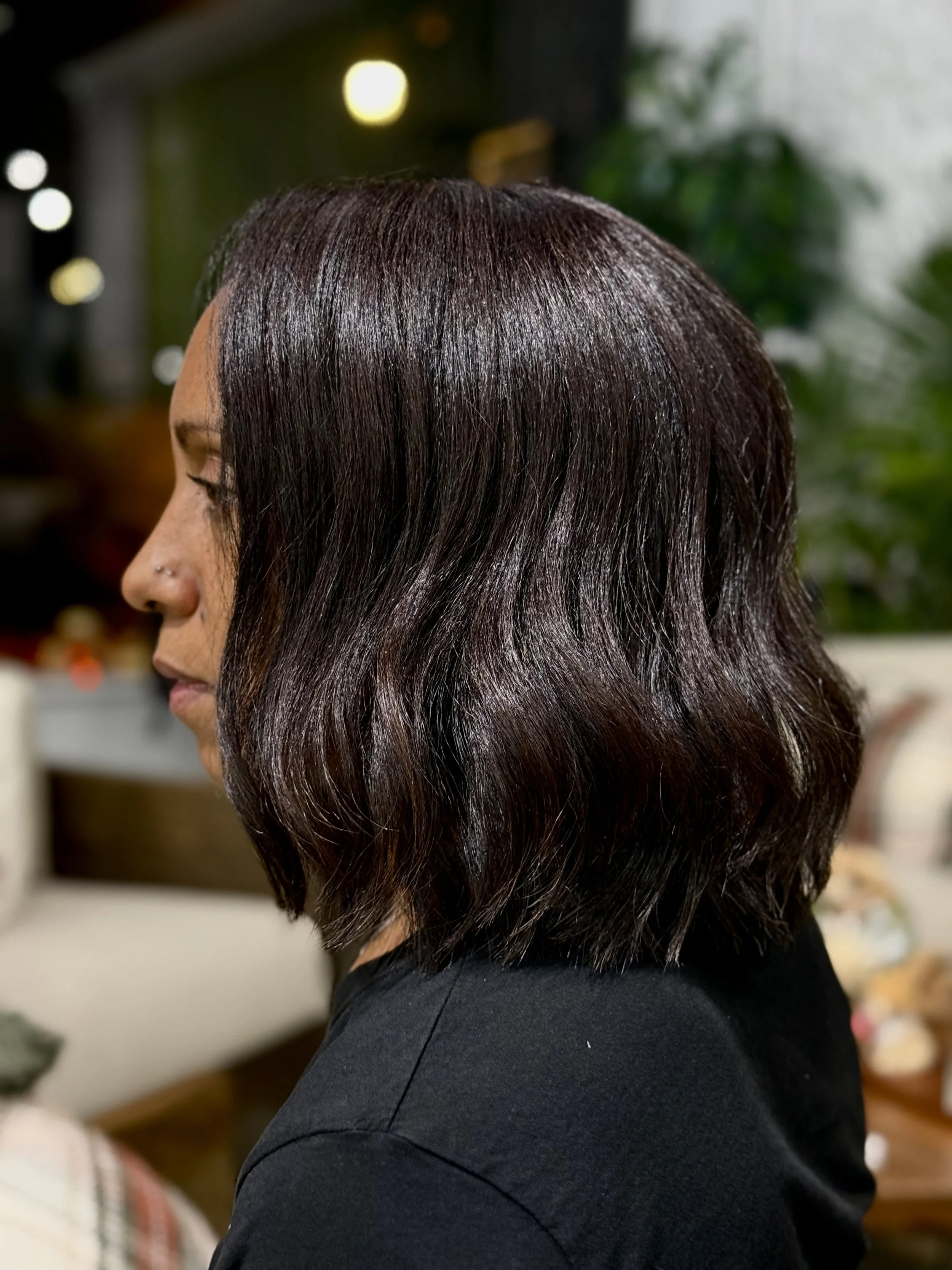 Side profile of a woman with shoulder-length, wavy, dark brown hair in a well-lit indoor setting.