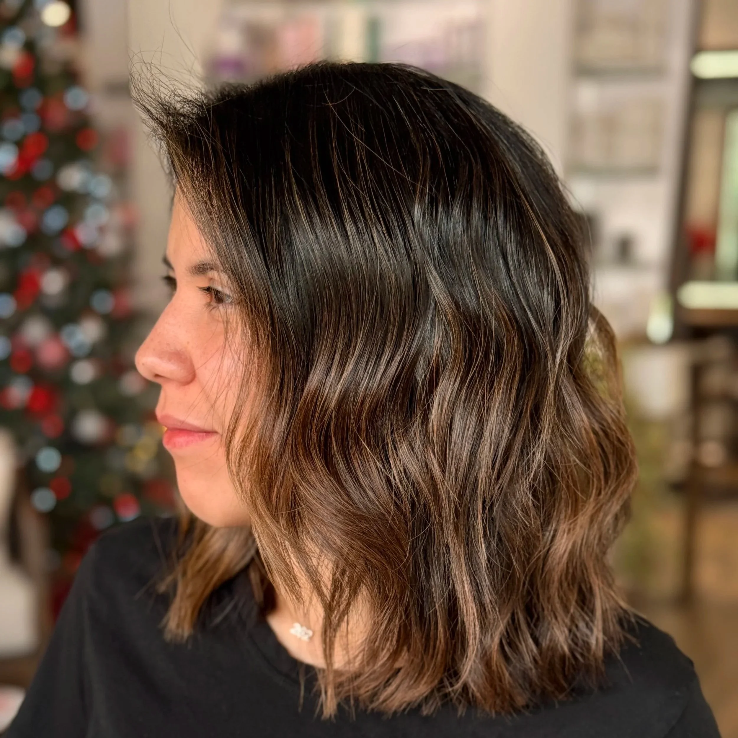 Profile of a woman with shoulder-length wavy brown hair with highlights, wearing a black top and a small flower-shaped necklace, standing indoors with a blurred background including a decorated Christmas tree.