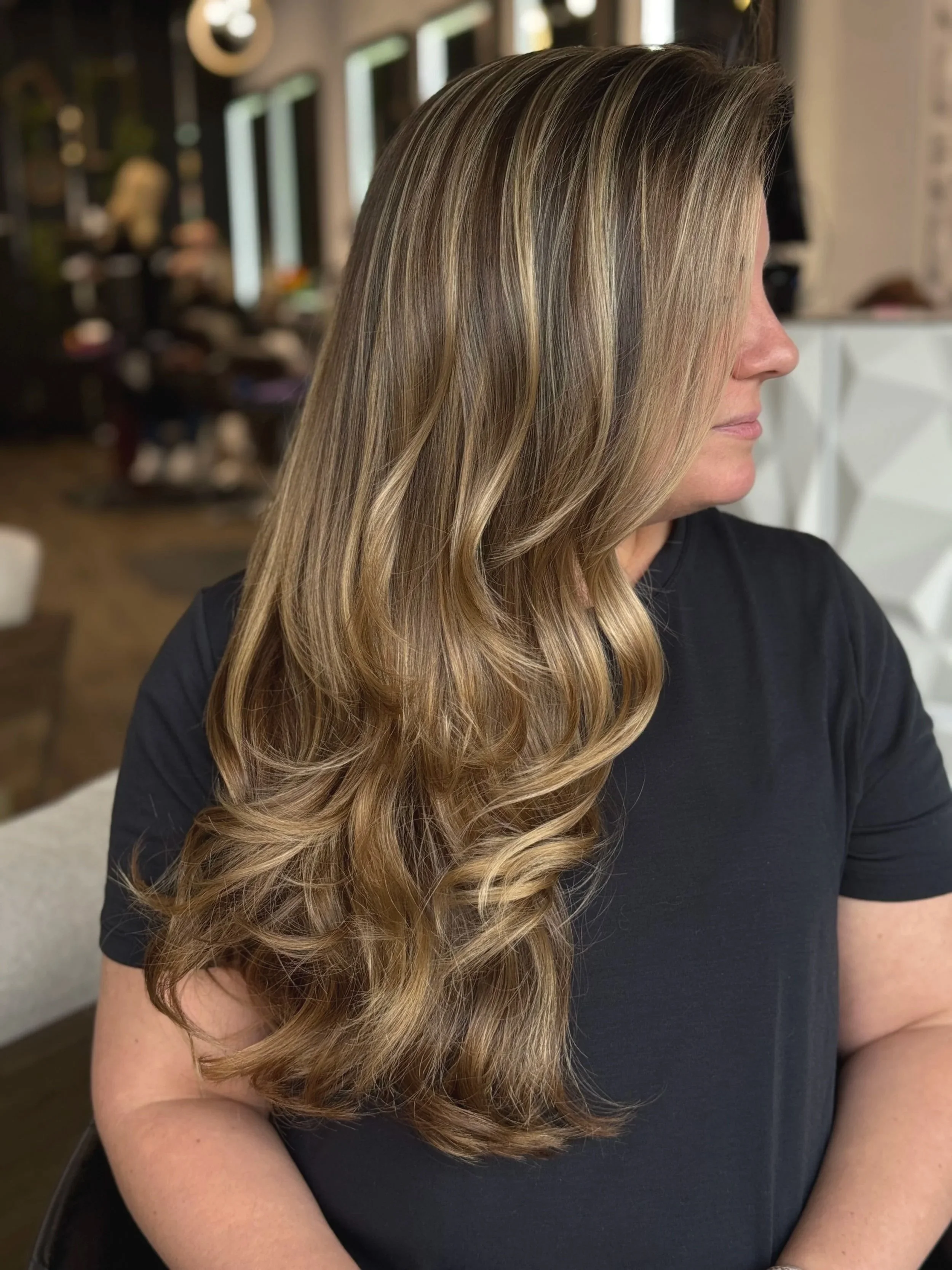 Side profile of a woman with long, styled wavy blonde hair, sitting in a salon with blurred background.
