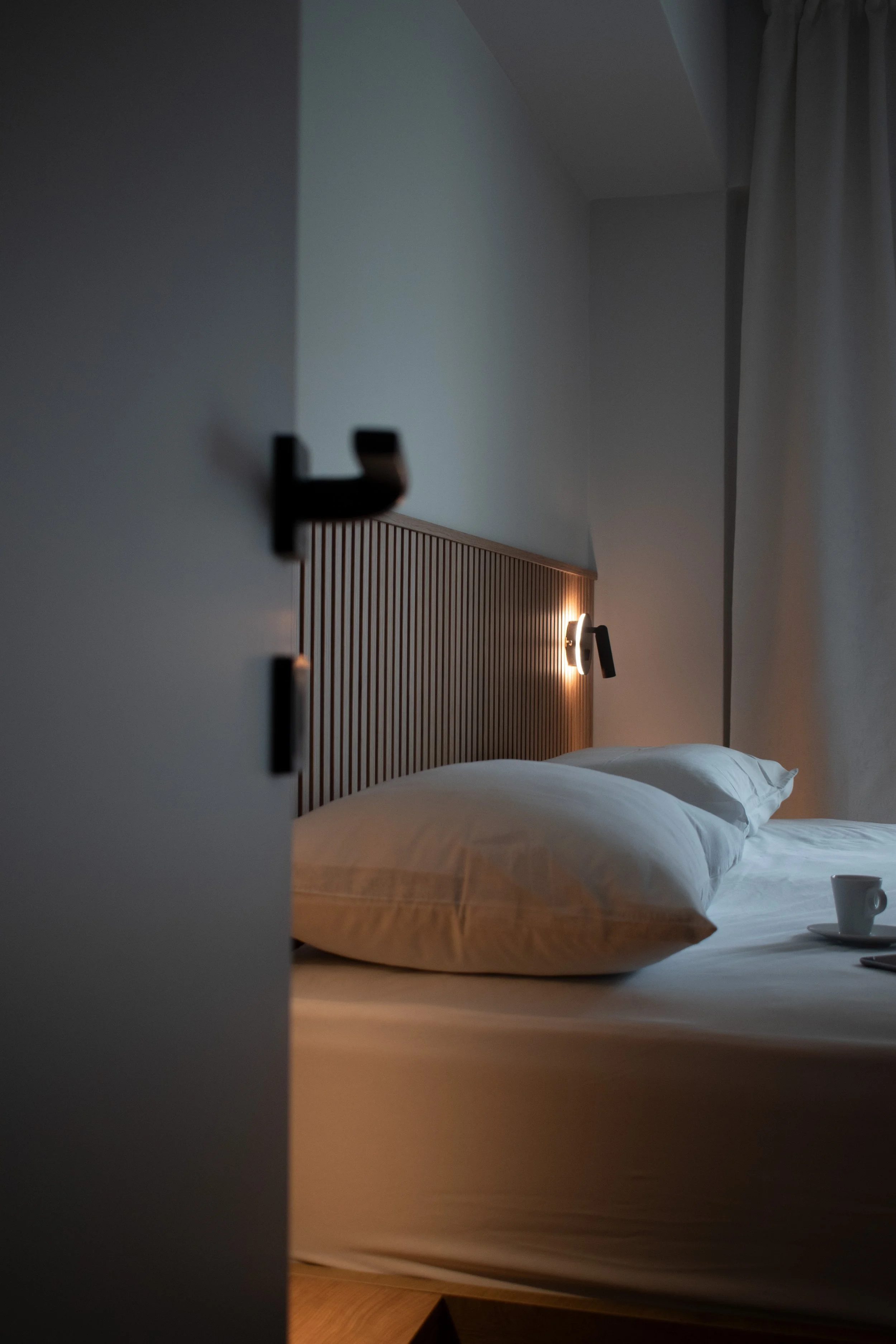 A hotel room with a bed, a wooden headboard, pillows, a bedside lamp, and a cup on a tray. The photo is taken through a partially open door, with curtains visible on the right side.