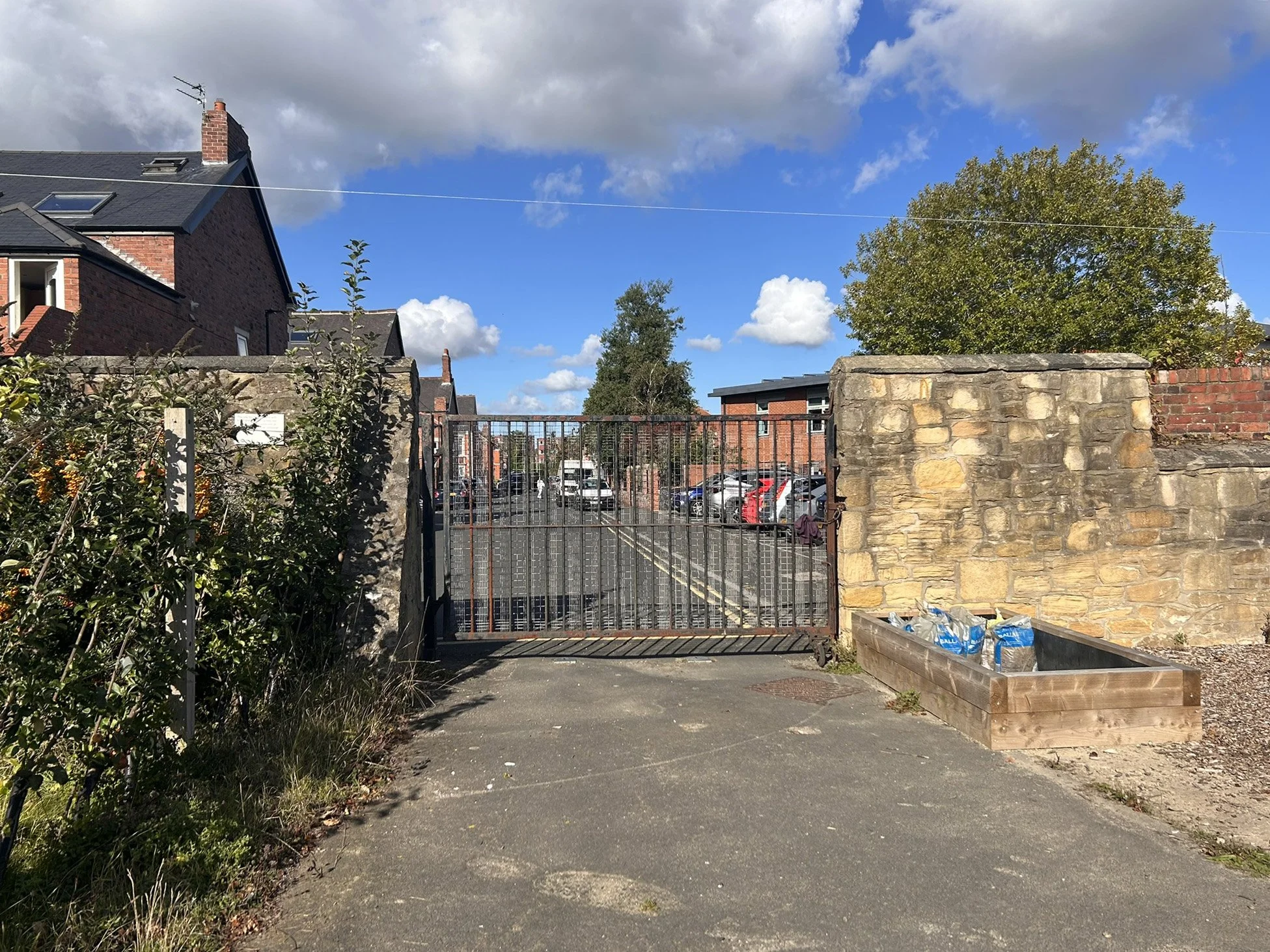 A gated entrance with a stone wall and metal gate leading to a parking lot filled with cars. On the right, there is a wooden raised garden bed with some bags of soil or mulch. The background includes a brick building, trees, and a blue sky with scatt
