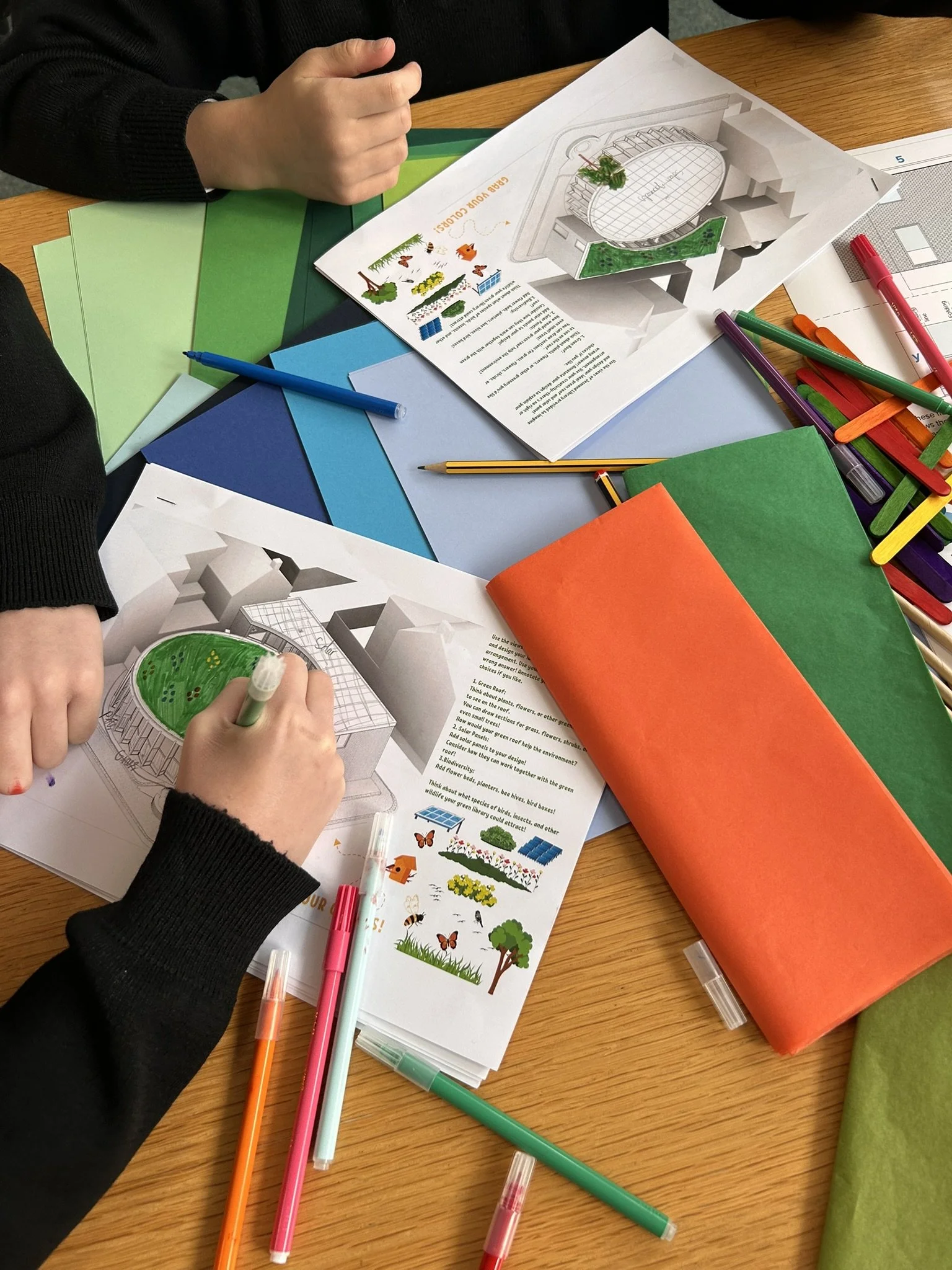 Hands of a person working on a detailed garden design plan, with colored markers, paper, and a printed illustration of a landscape on a wooden table.