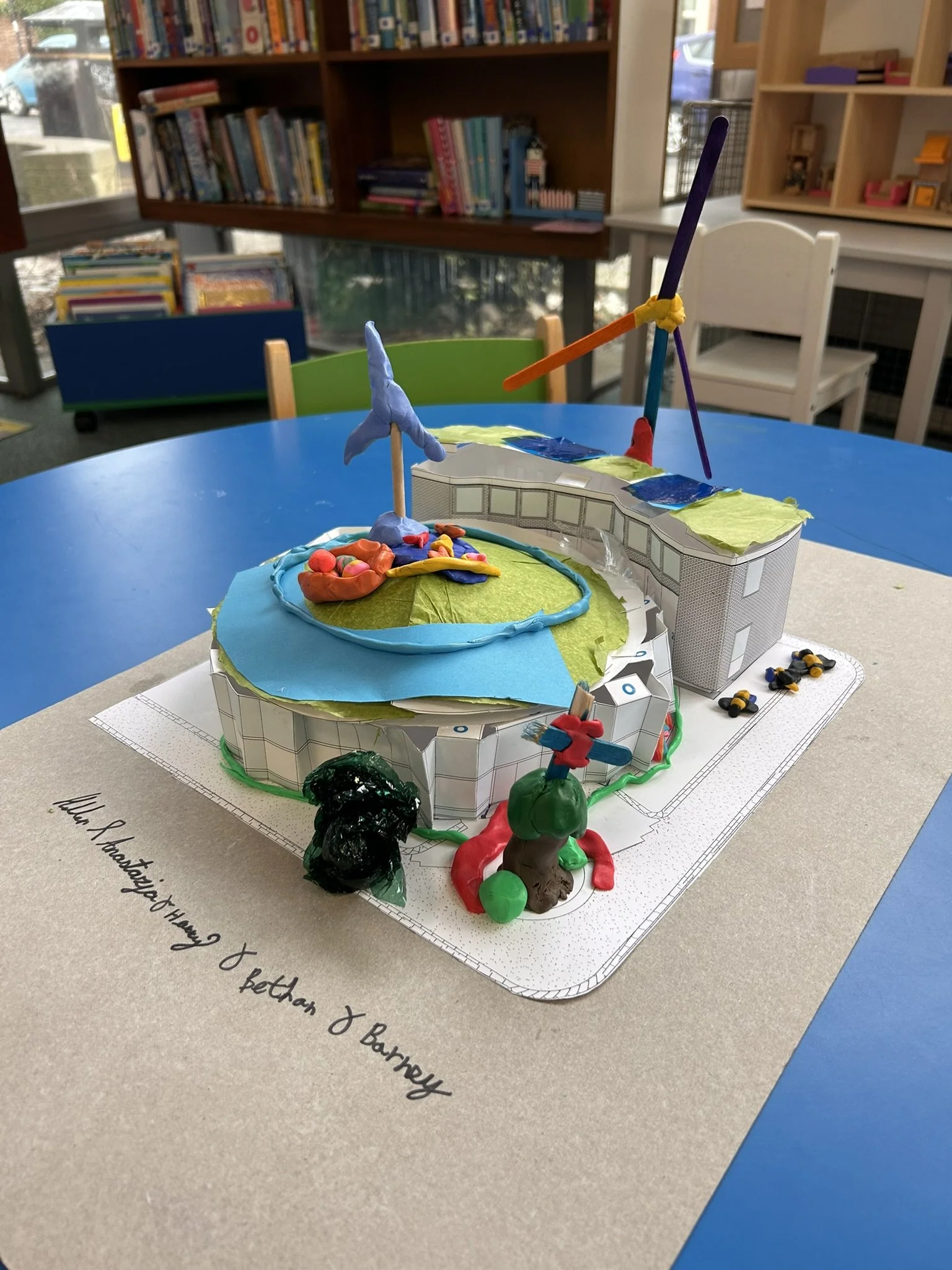 Colorful handmade architectural model with a circular design, featuring a spinning wind turbine and trees, set on a paper base with a handwritten note.
