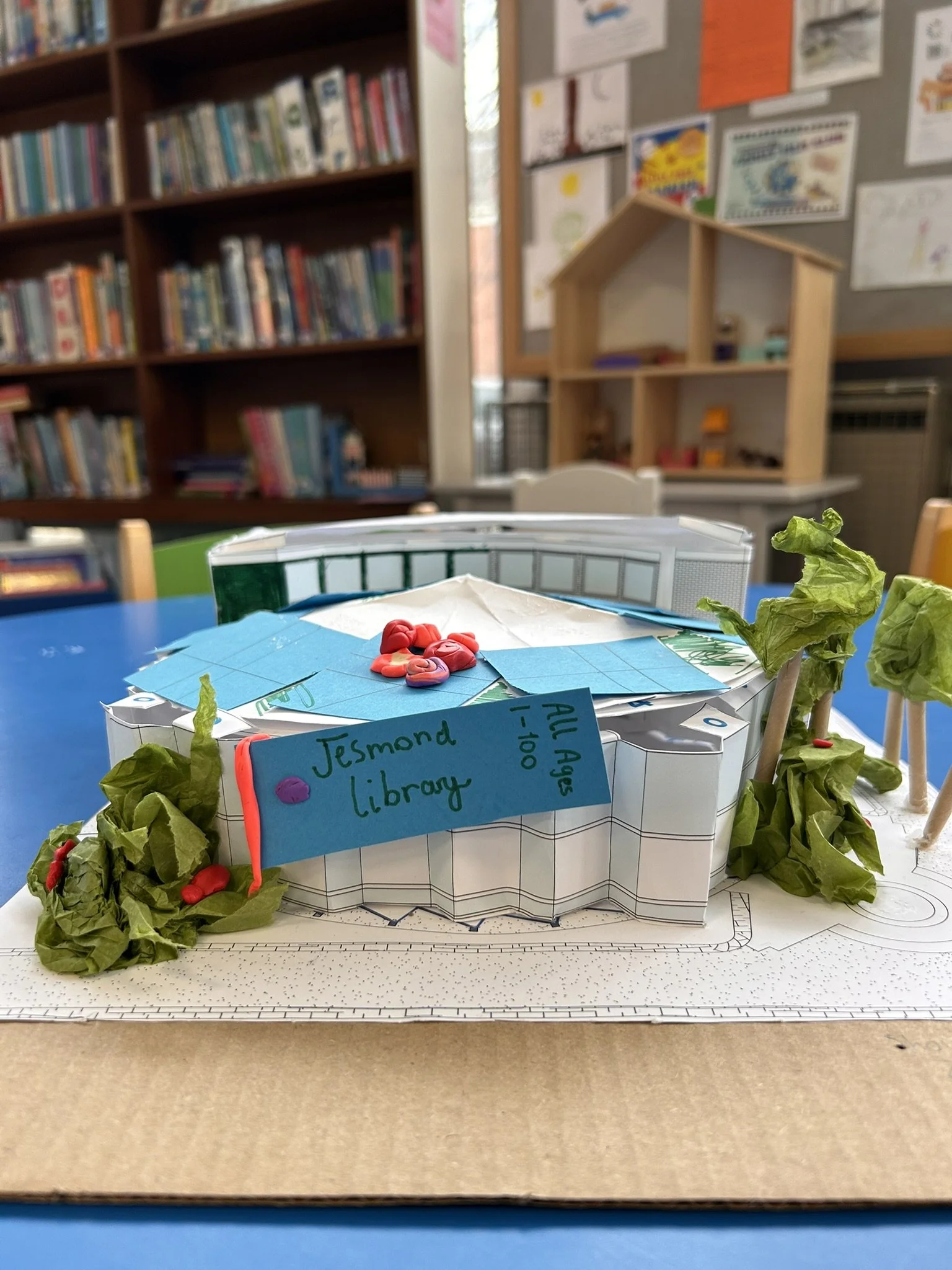 A paper craft model of a library with green crumpled paper trees on each side, a blue paper roof, and a blue sign that reads 'Jesmond Library' in handwritten text. The background shows a classroom with bookshelves and bulletin boards.