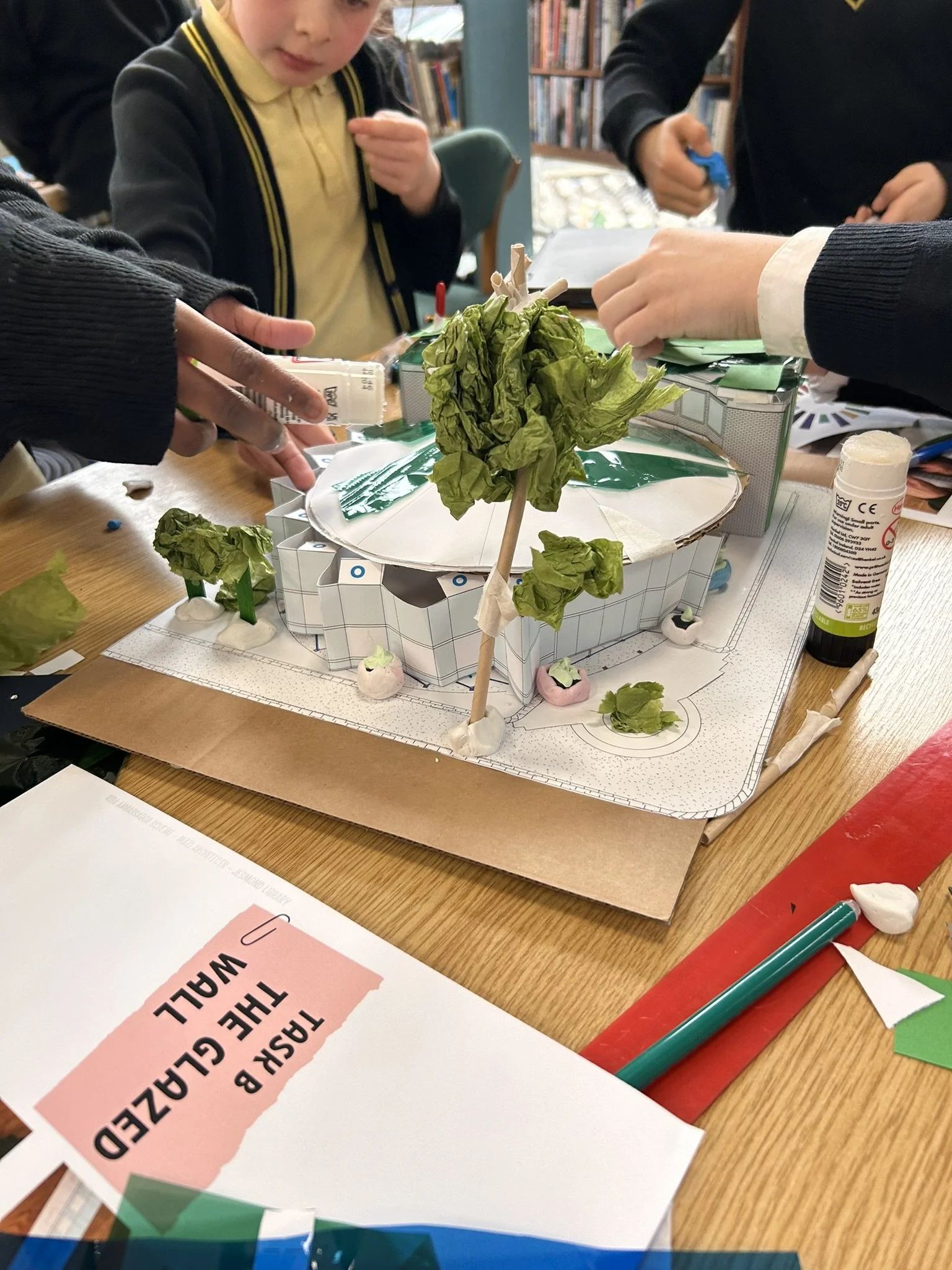 Children working on a craft project with a model of a building, paper, glue, and markers on a wooden table.