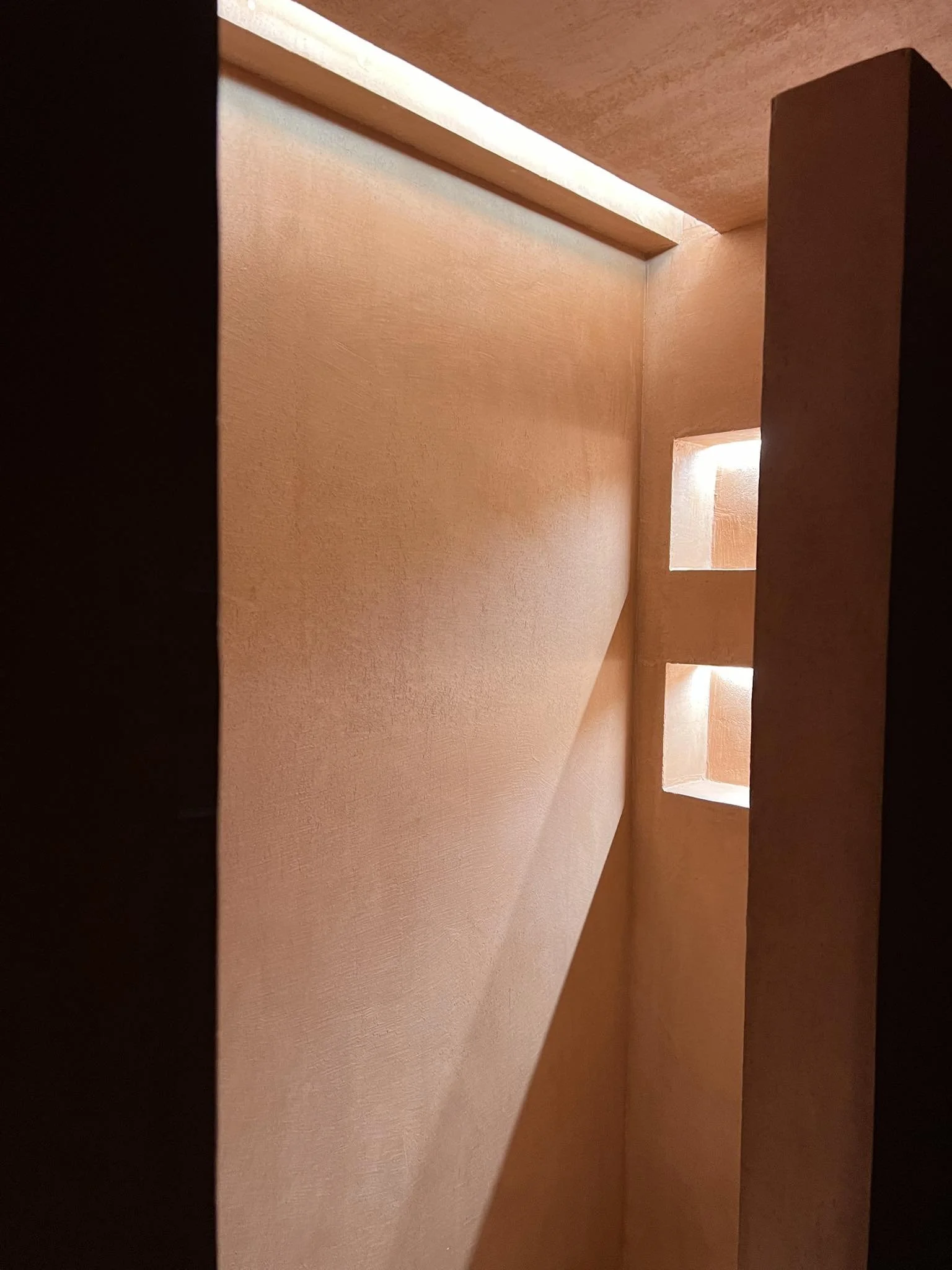 A view of an interior corner with a cut-out window near the ceiling, illuminated by natural light, featuring warm-colored walls and a ceiling.