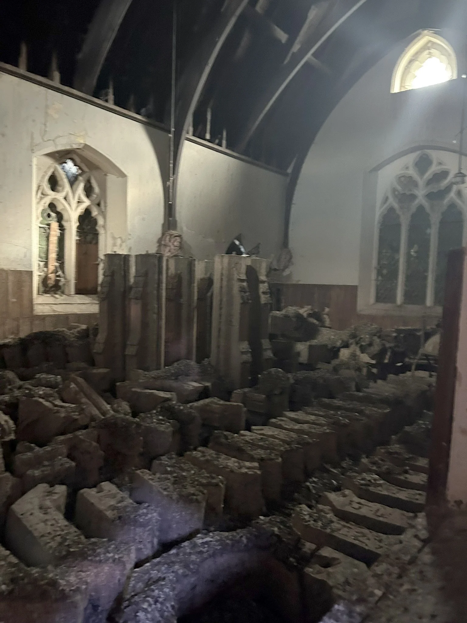 The interior of a church under construction or renovation, with exposed stone and brick walls, rubble on the floor, and a partially finished altar area. Light comes through a stained glass window.
