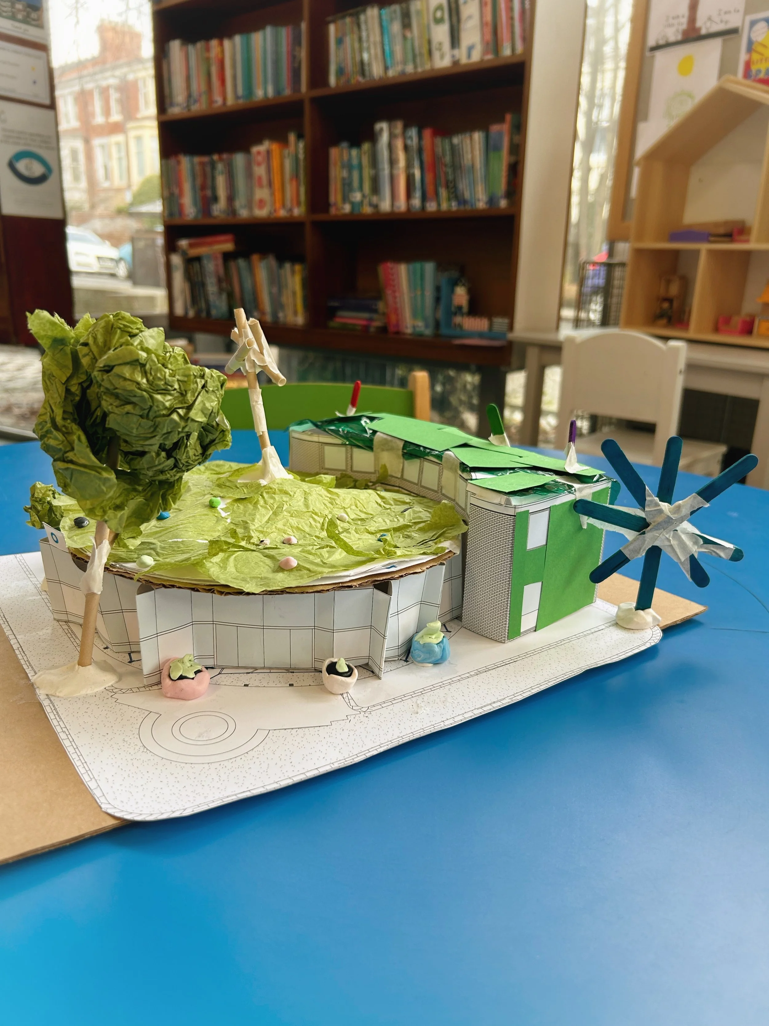 A colorful, handmade diorama of a modern house with trees and landscaping, made from paper and other craft materials, placed on a table with a blueprint background. In the background, a bookshelf filled with books and a window can be seen.