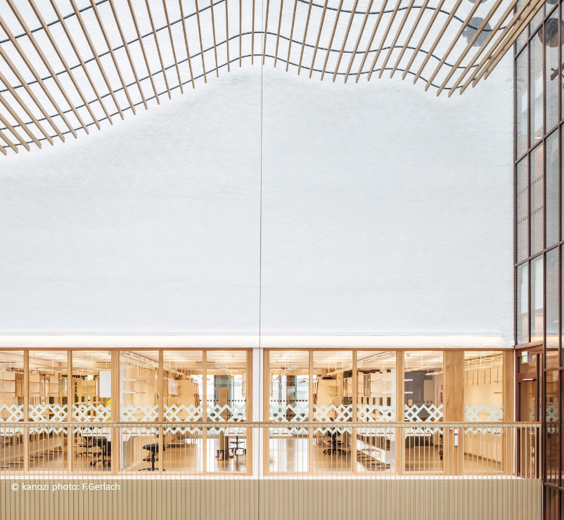 Modern interior view of a building with a large open space, wooden ceiling slats, glass walls, and a visible office area behind panels.