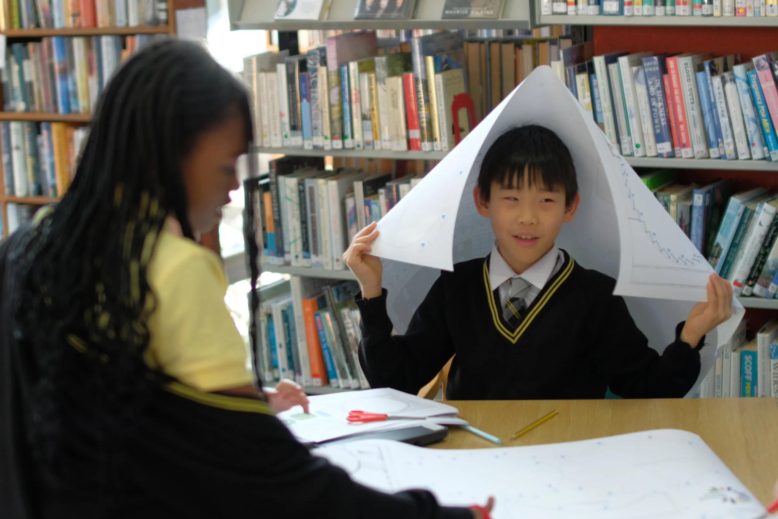 A boy in a school uniform holding a large paper sculpture of a mountain with snow-capped peaks, inside a library with shelves of books. A girl with braided hair and a yellow top is in the foreground, slightly out of focus.