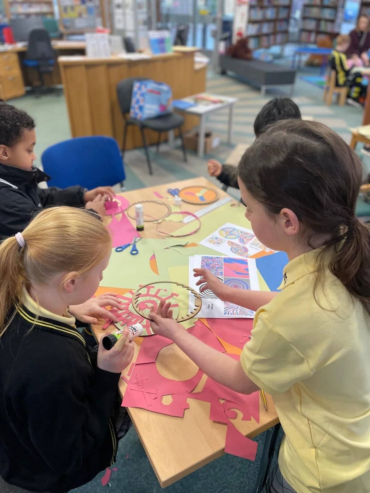 Children engaging in arts and crafts activities at a table in a library. They are cutting paper, using glue, and creating designs with colored paper and printed patterns.