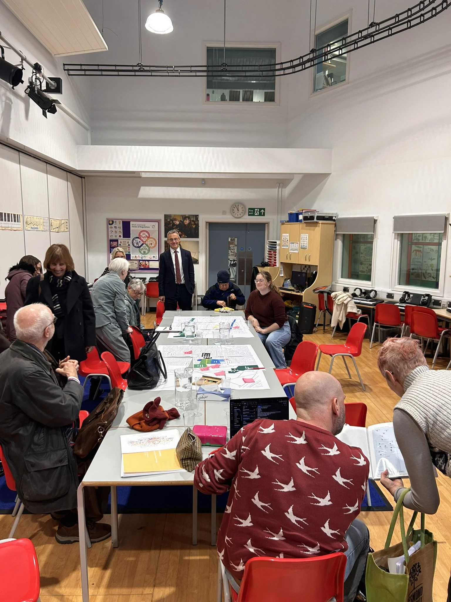 A group of people gathered in a room around a table, some sitting and others standing, engaged in conversation and activities. The room has white walls, large windows, and various office supplies and equipment.