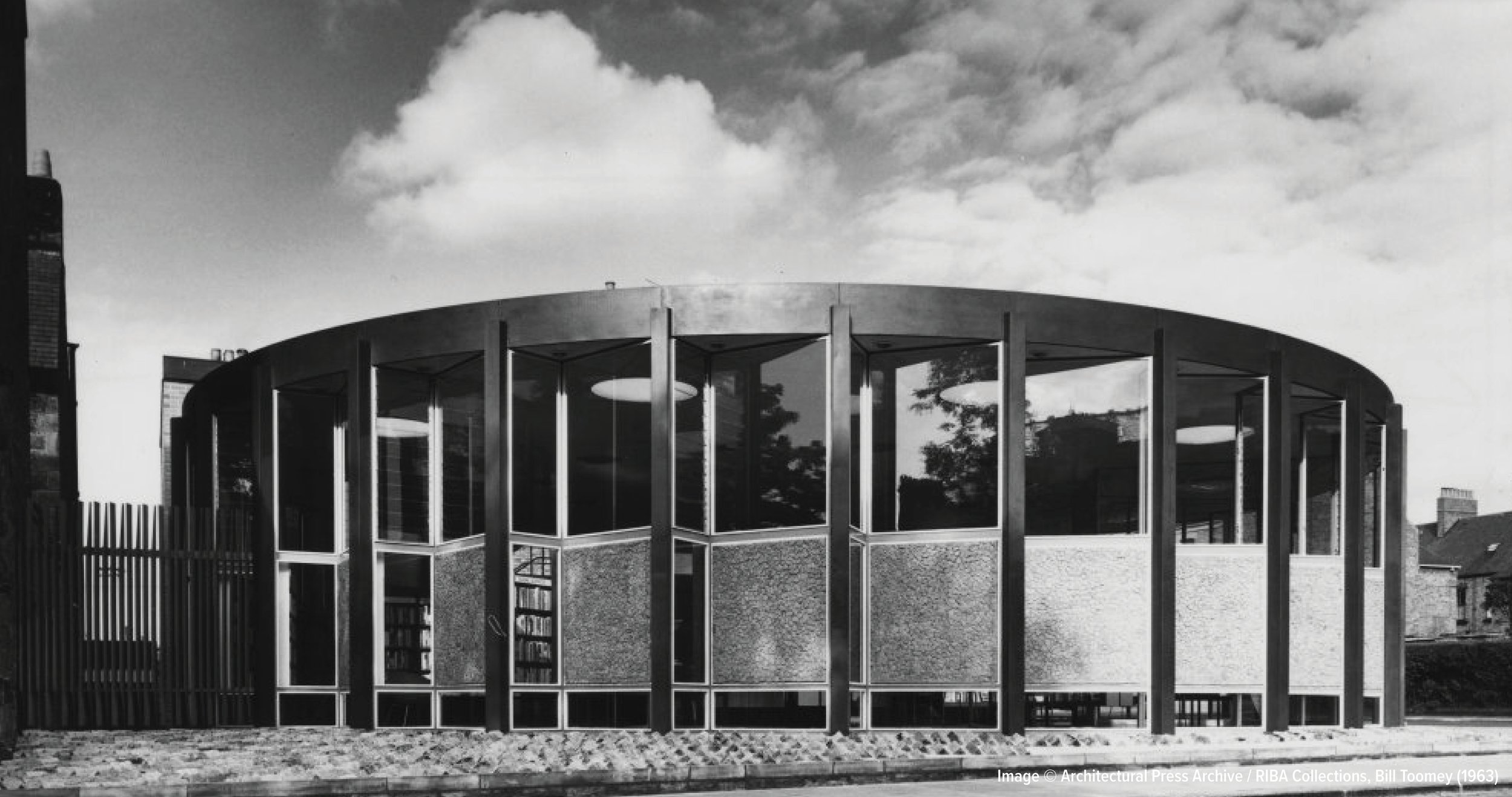 Black and white photo of a modern round building with large windows and vertical structural supports, under a cloudy sky.