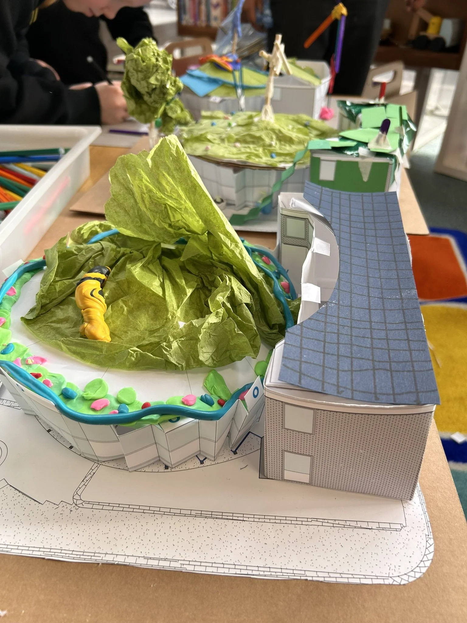 A detailed architectural model of a building with several sections and landscaping elements, placed on a table with art supplies and people working in the background.