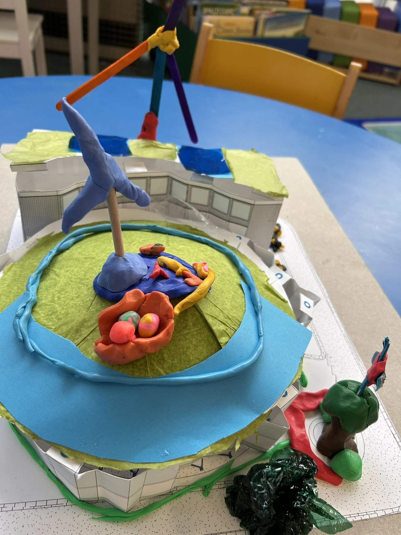 A colorful model of a sports stadium and field made from various craft materials and paper, placed on a table in a classroom.