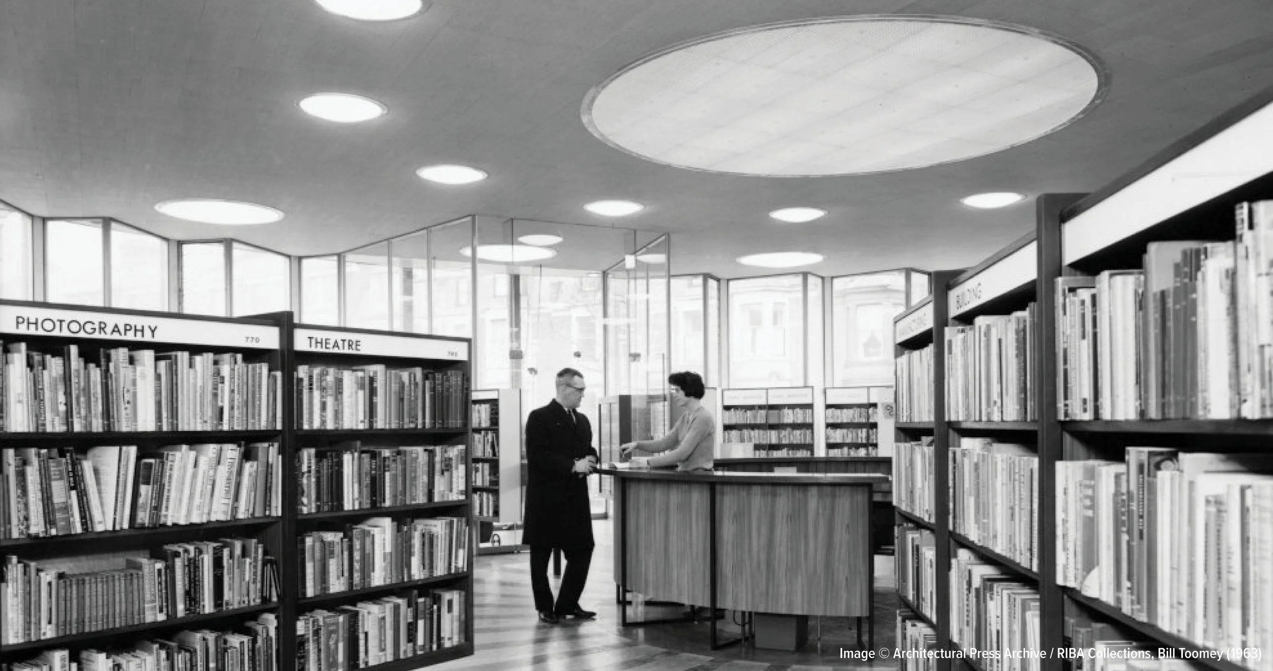 Black and white photo of a bookstore or library interior with bookshelves labeled 'Photography' and 'Theatre'. Two people are engaged in conversation near a counter. Large windows in the background let in natural light. The ceiling has circular reces