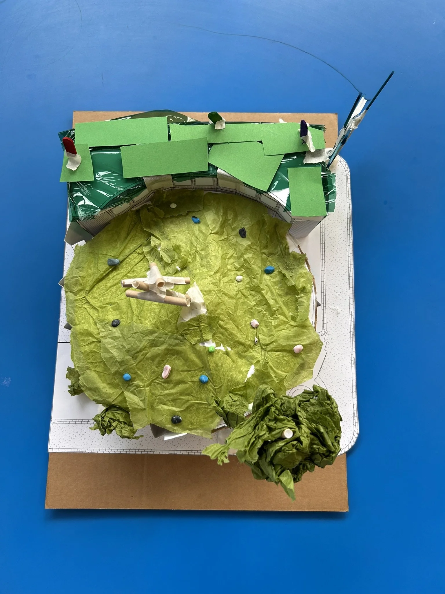 A miniature model of a landscape or cityscape made from paper and other craft materials, featuring green paper representing grass or trees, with some small poles and a paper structure resembling a bench or barrier, and a small leafy bush at the botto