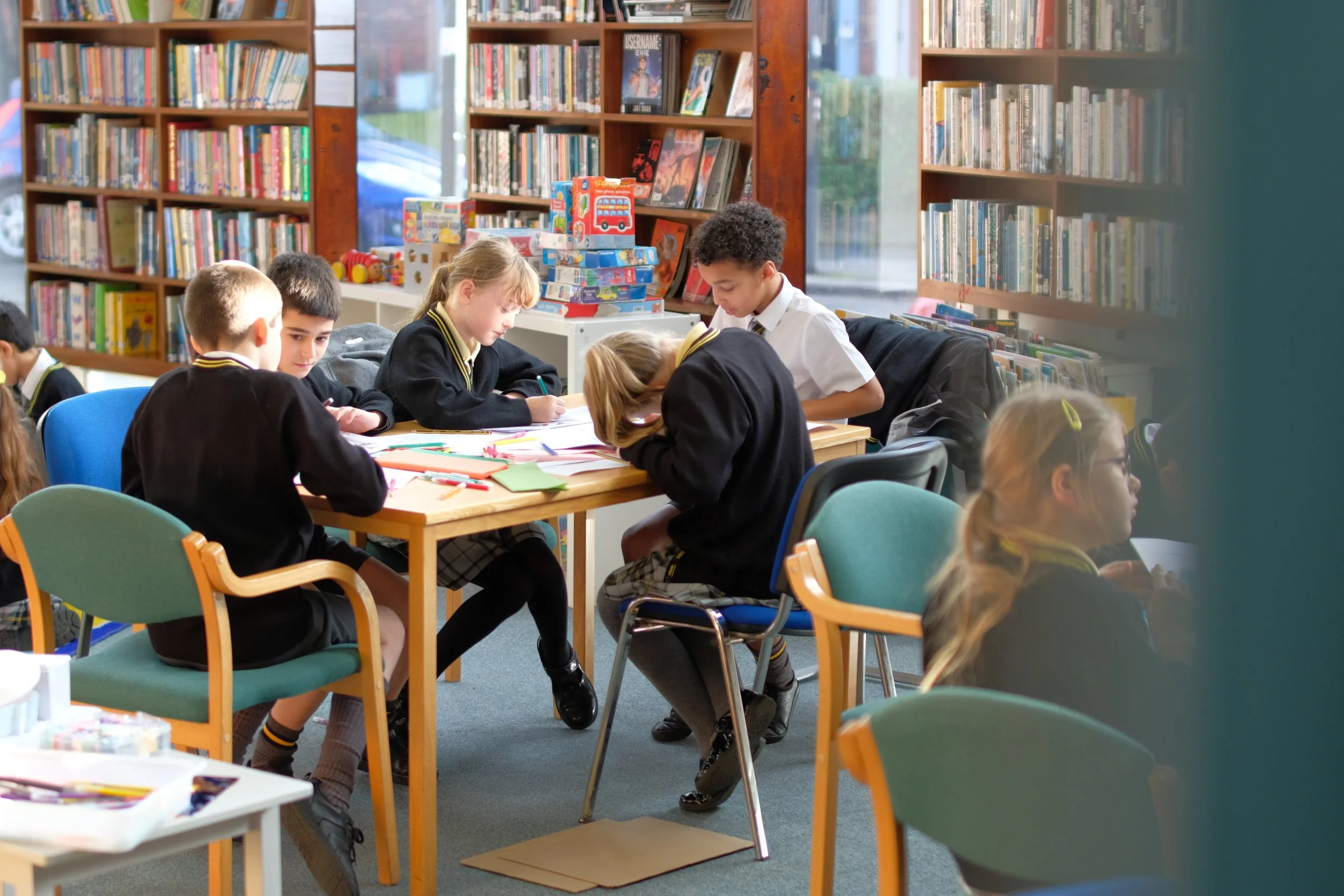 Children sitting around tables with art supplies in a library or bookstore, surrounded by shelves of books.