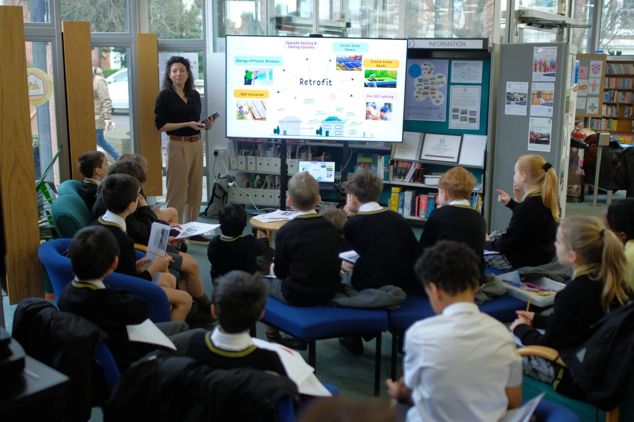 A classroom presentation where a woman is pointing at a large screen displaying a diagram about retrofit processes. Several students in school uniforms are seated and attentively listening, with some taking notes.