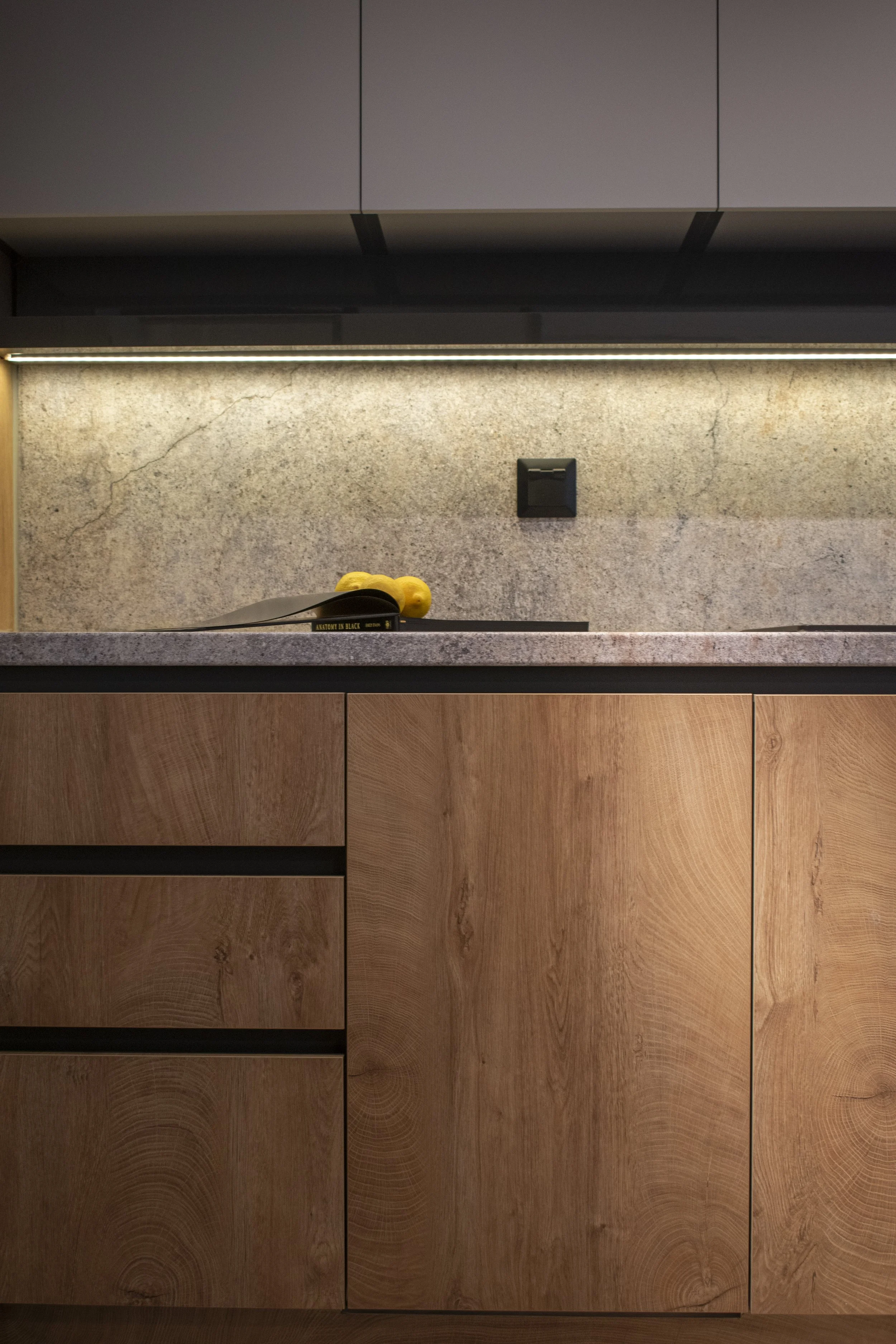 Kitchen countertop with a black spatula and a yellow rubber glove resting on a cookbook titled 'Anatomy in Black', with a textured beige backsplash and wooden cabinets below.