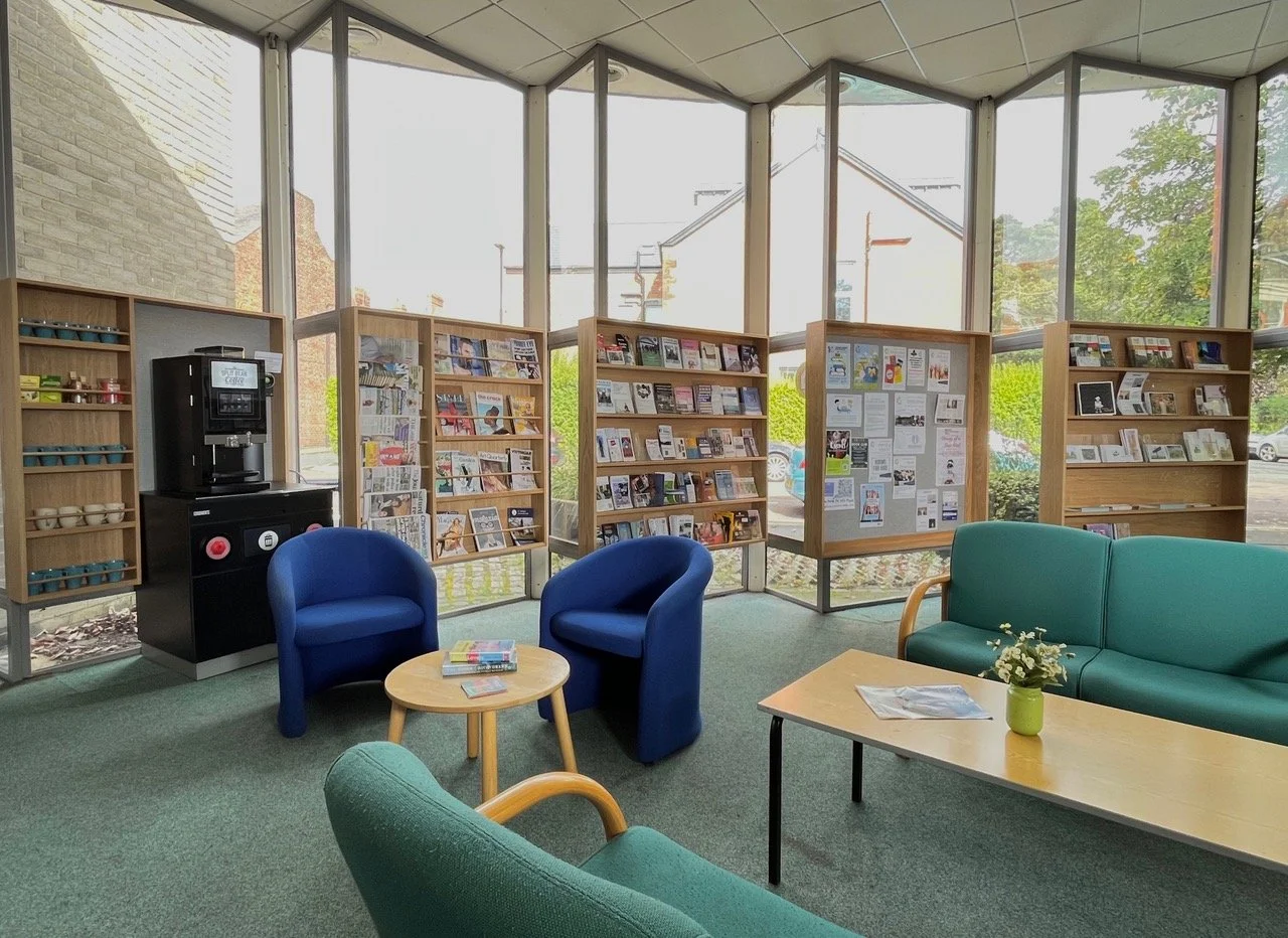A cozy reading corner in a library or community space with large windows, theater seating, bookshelves, a coffee machine, magazines, and a small table with a flower vase.