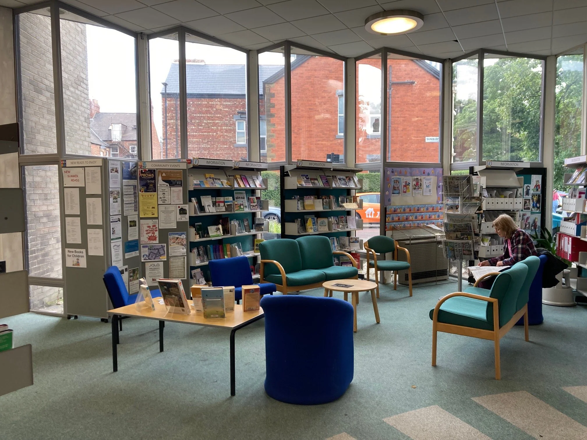 Inside a library with large windows showing brick buildings outside. The room has chairs and bookshelves with books and pamphlets. A woman is seated reading a newspaper.