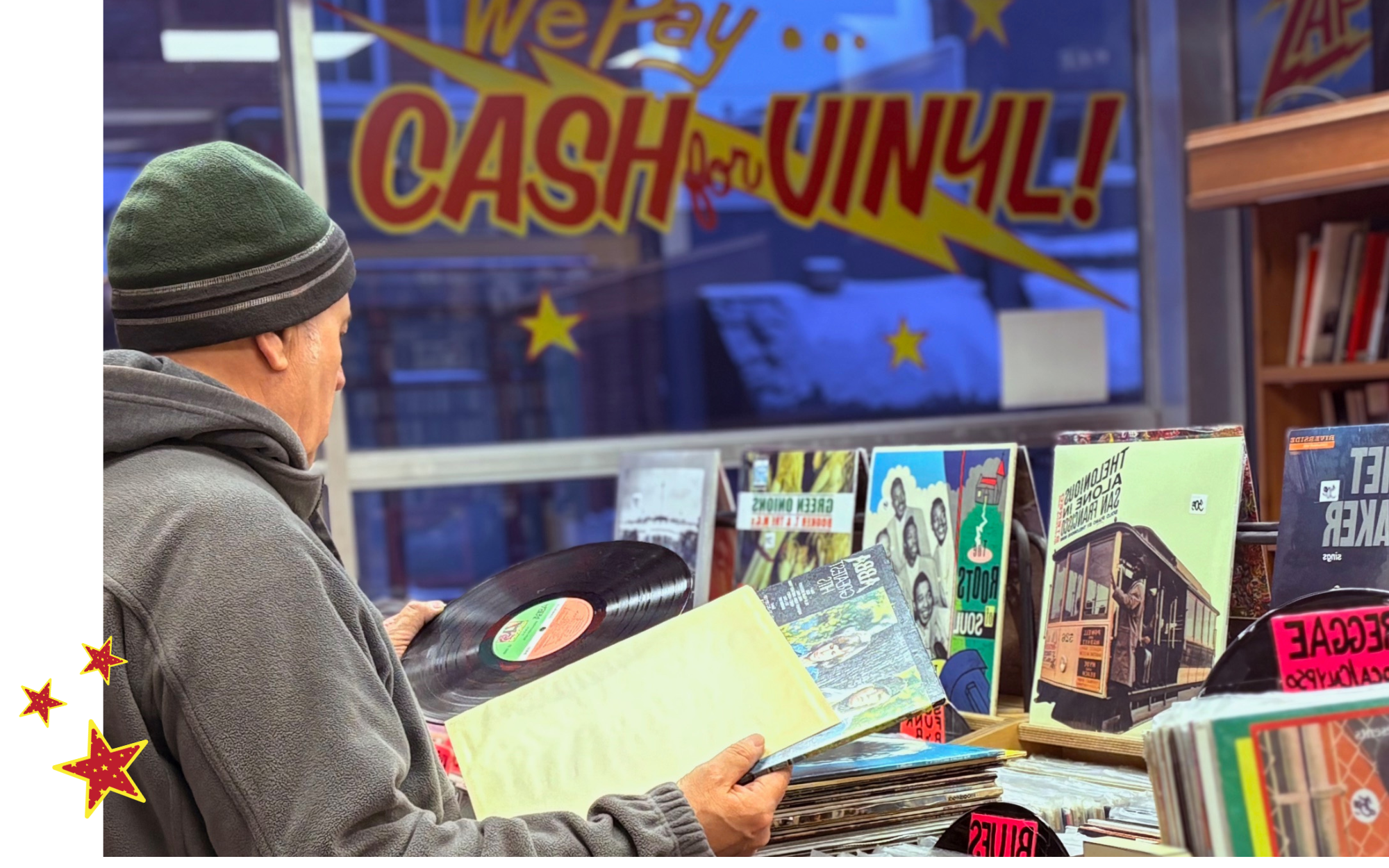 A man in a gray coat and beanie looking at vinyl records in a store, with a large colorful sign in the background that says 'We Pay CASH for VINYL!'.