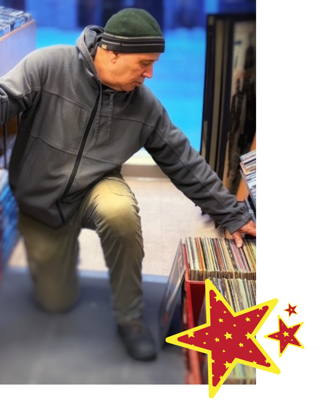A man wearing a green beanie and a gray hoodie is kneeling and browsing through a collection of records in a store.