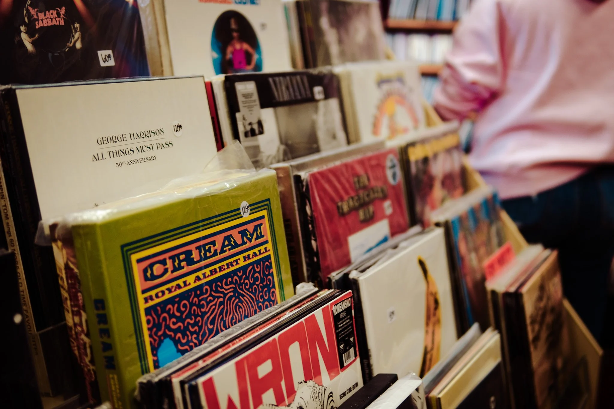 A section of vinyl records on display in a record store, including albums by George Harrison, The Beatles, and Black Sabbath, with a person in a pink shirt partially visible on the right.