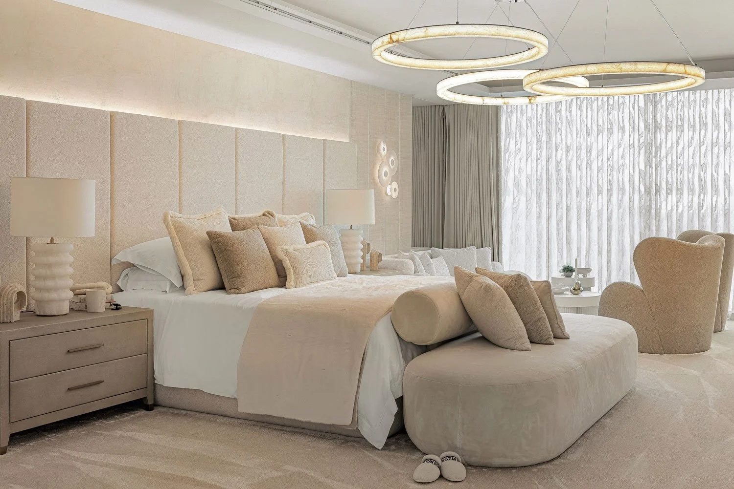 A luxurious bedroom with a large bed adorned with beige and cream pillows, two bedside lamps, a plush circular ottoman at the foot of the bed, and a sitting area with armchairs by a large window with sheer curtains and beige drapes. Modern circular c