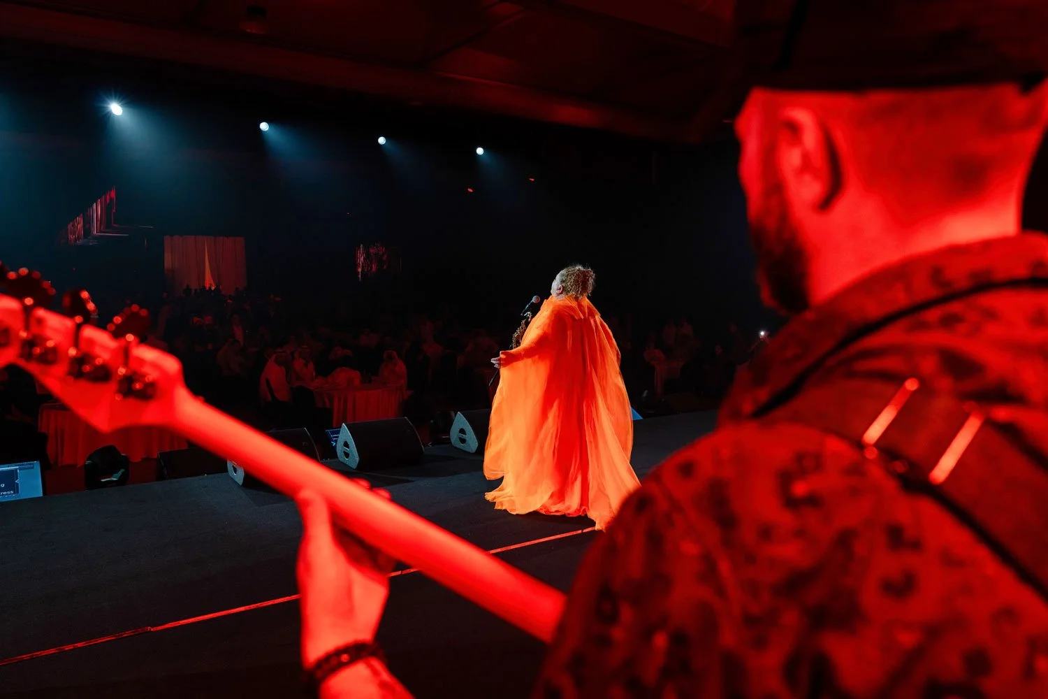 A performer wearing a flowing orange dress singing on stage with a microphone, seen from backstage perspective, with the audience in the background and a guitarist in the foreground.