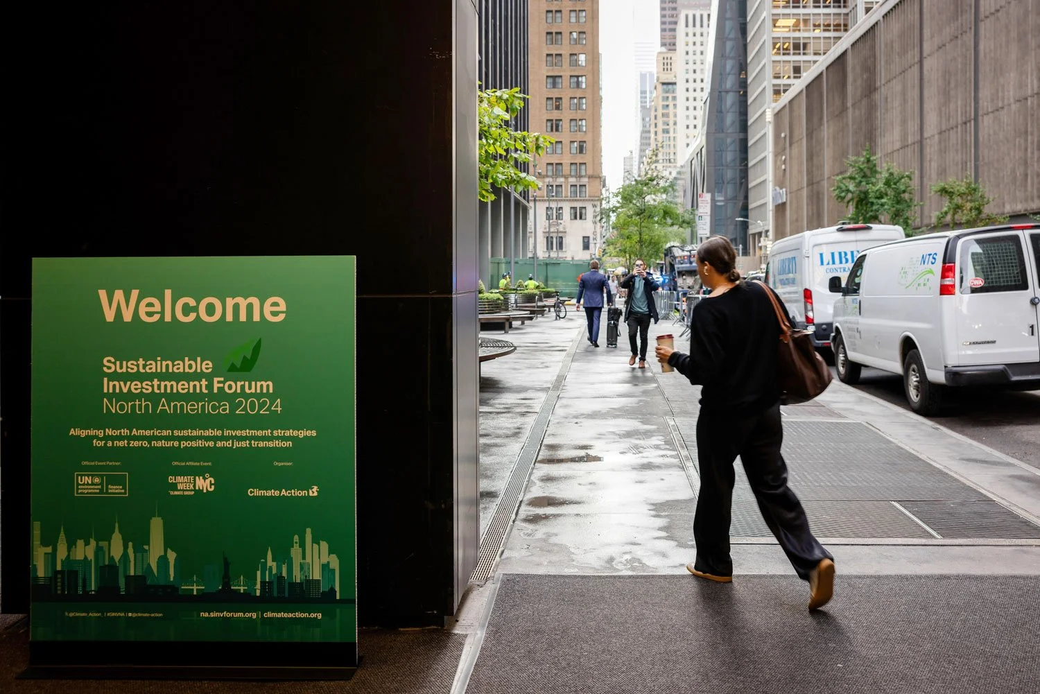 A sidewalk scene outside a conference venue with a large green sign that reads 'Welcome Sustainable Investment Forum North America 2024' and an urban street with pedestrians and parked vans.