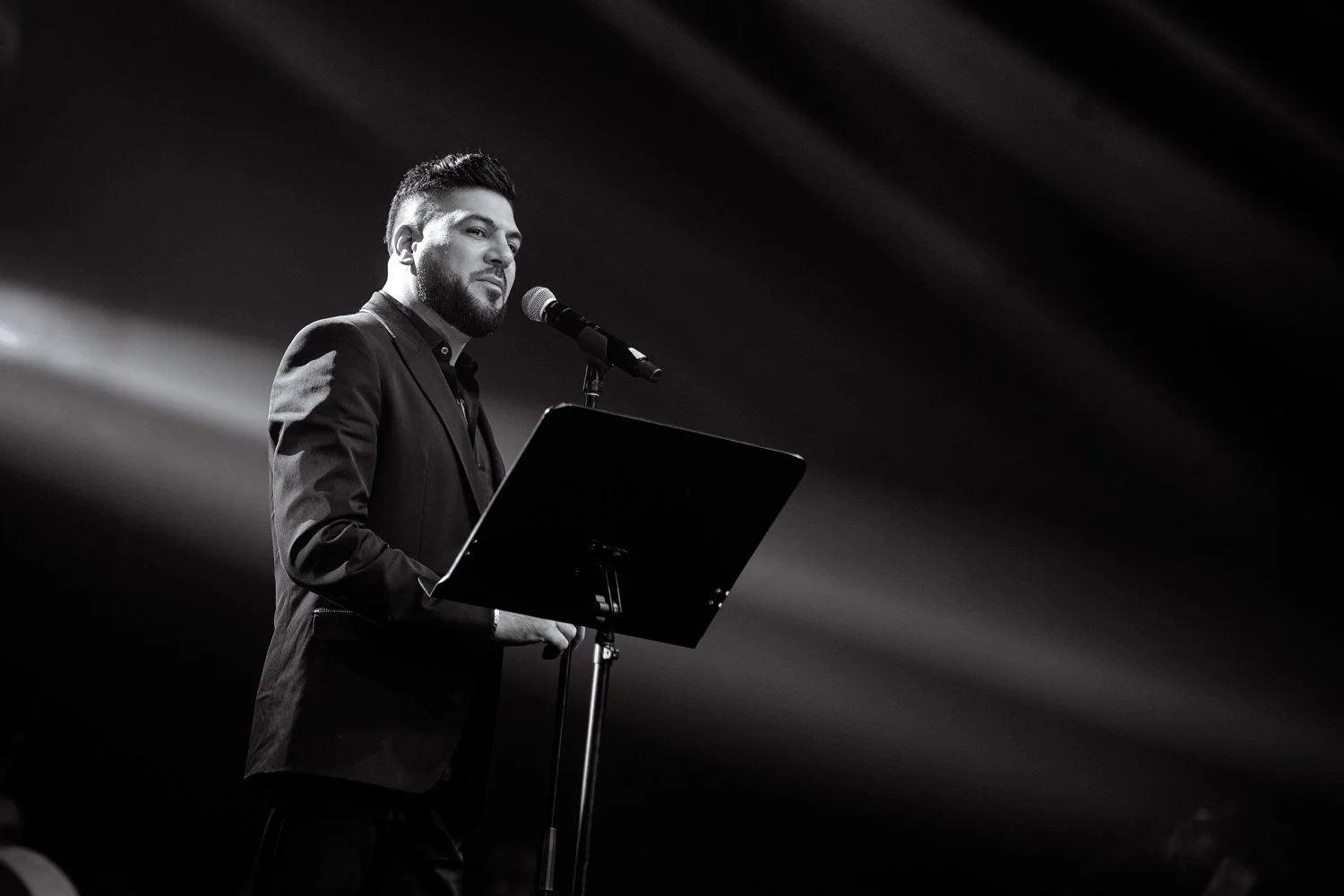 A man with a beard and short hair, dressed in a suit, standing at a microphone on a stand with a music stand, speaking or singing on a stage with dramatic lighting.