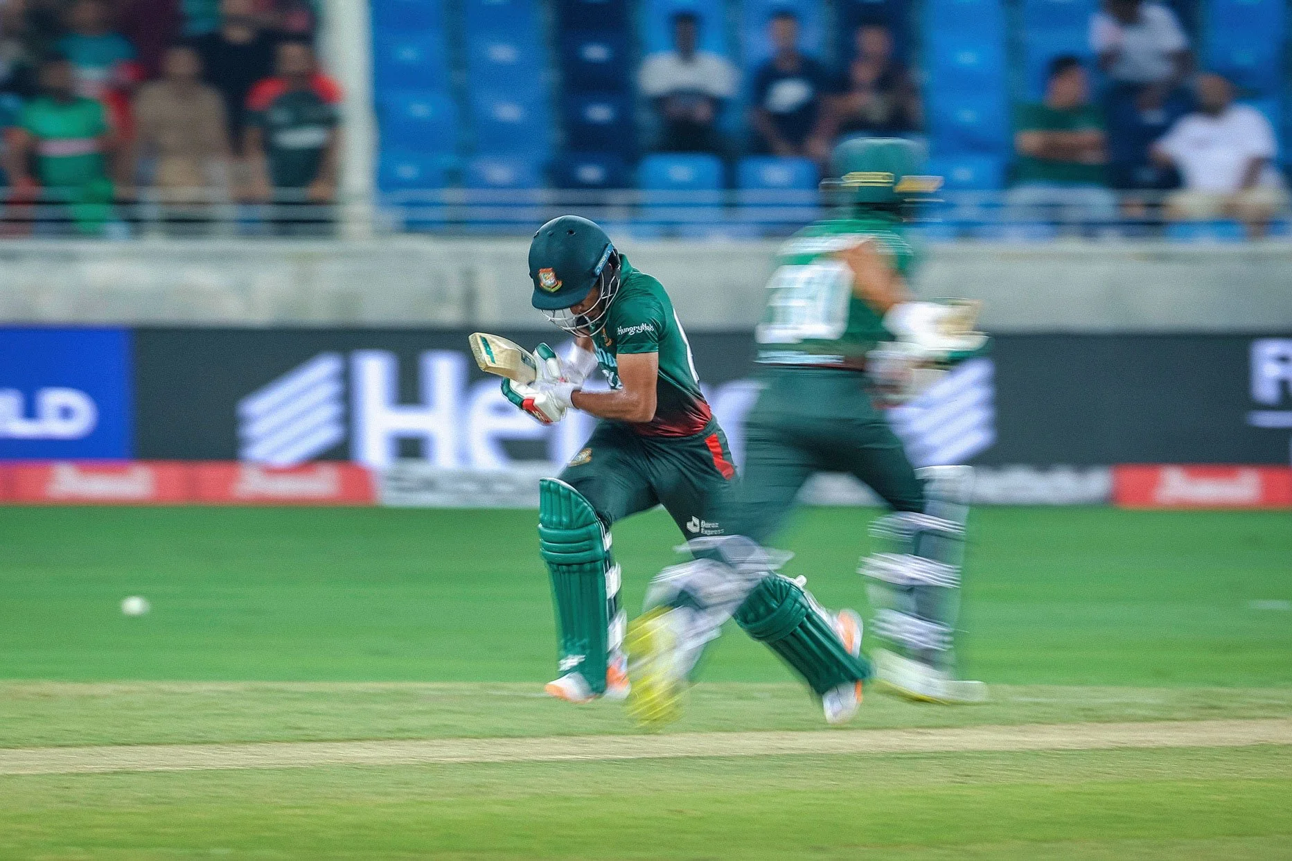 Cricket player running on field in green uniform with batting gear, blurred background of spectators.