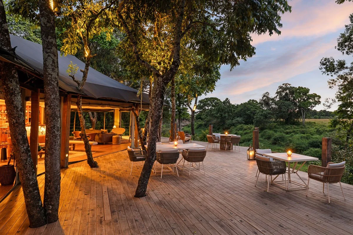 An outdoor deck with tables and chairs, lit by lanterns, surrounded by trees and greenery at sunset.